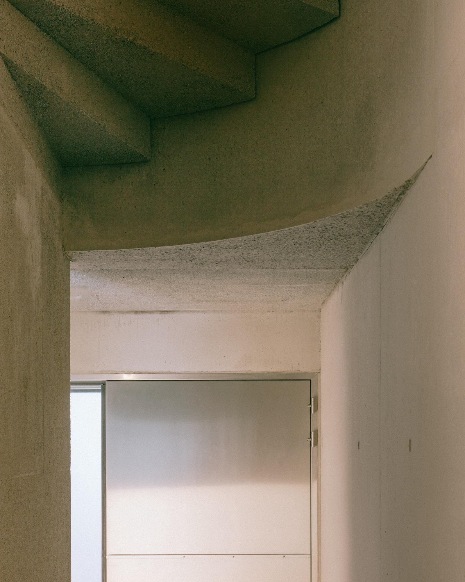 Xavier Hufkens Gallery by Robbrecht en Daem Architecten, photographed by Jeremy Piret — Underside of concrete stair showing curved soffit, raw aggregate texture with subtle formwork grain, doorway to lit gallery beyond.