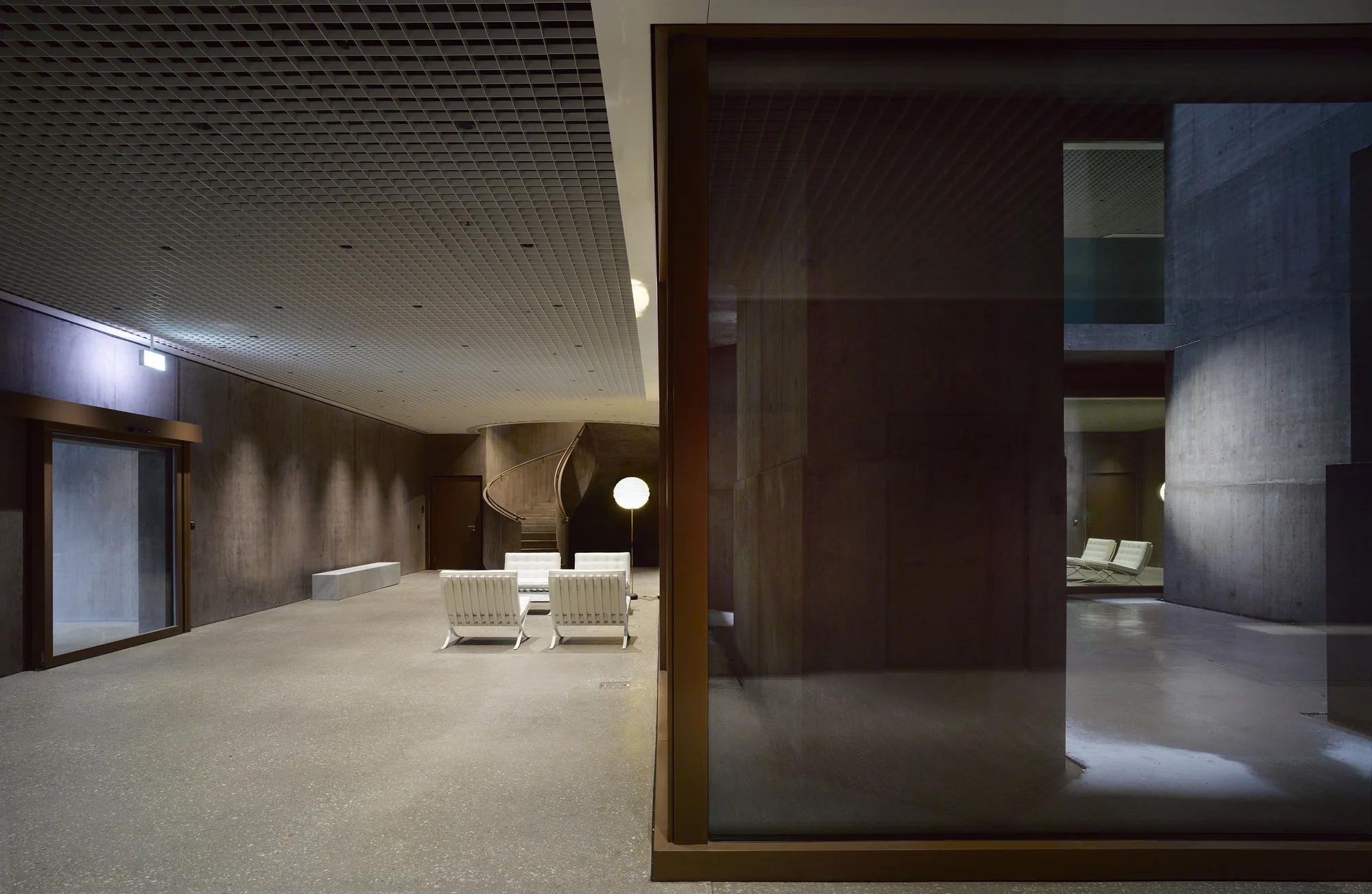 Baloise Insurance Company Office Tower by Valerio Olgiati, photographed by Bas Princen — Lobby space with Barcelona chairs in white leather faces a curved concrete stair, bronze-framed glass enclosures dividing the volume.