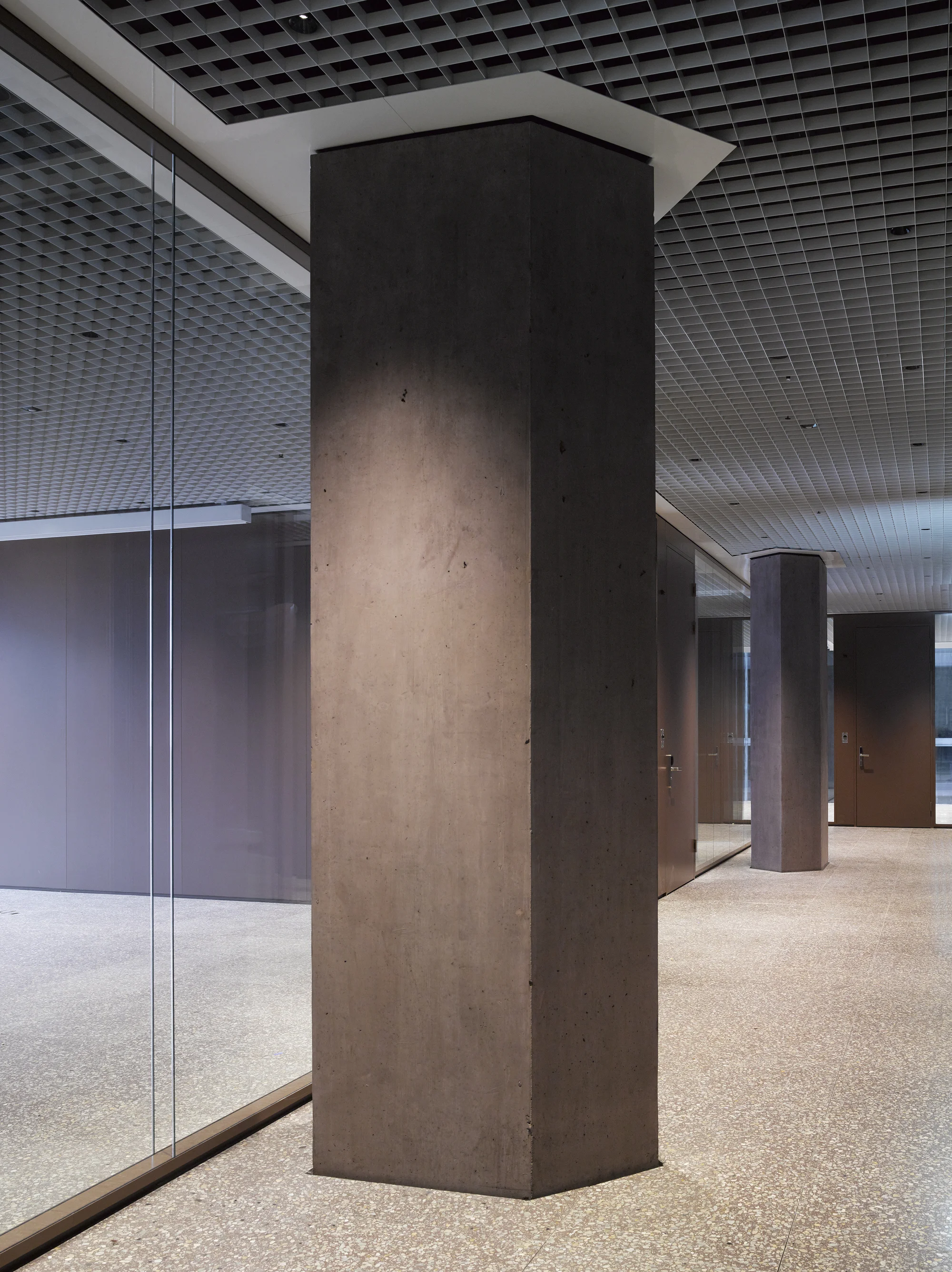 Baloise Insurance Company Office Tower by Valerio Olgiati, photographed by Bas Princen — Interior lobby column in smooth brown concrete meets the terrazzo floor, an egg-crate metal ceiling above and glass partitions beyond.
