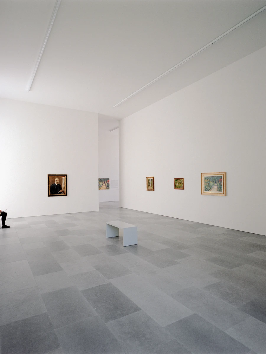Bastian Gallery by John Pawson, photographed by Julian Holzwarth — Interior of the main gallery room: grey stone tile floor, white plaster walls hung with gilt-framed canvases at measured intervals, a single white bench at centre, a figure seated at the…