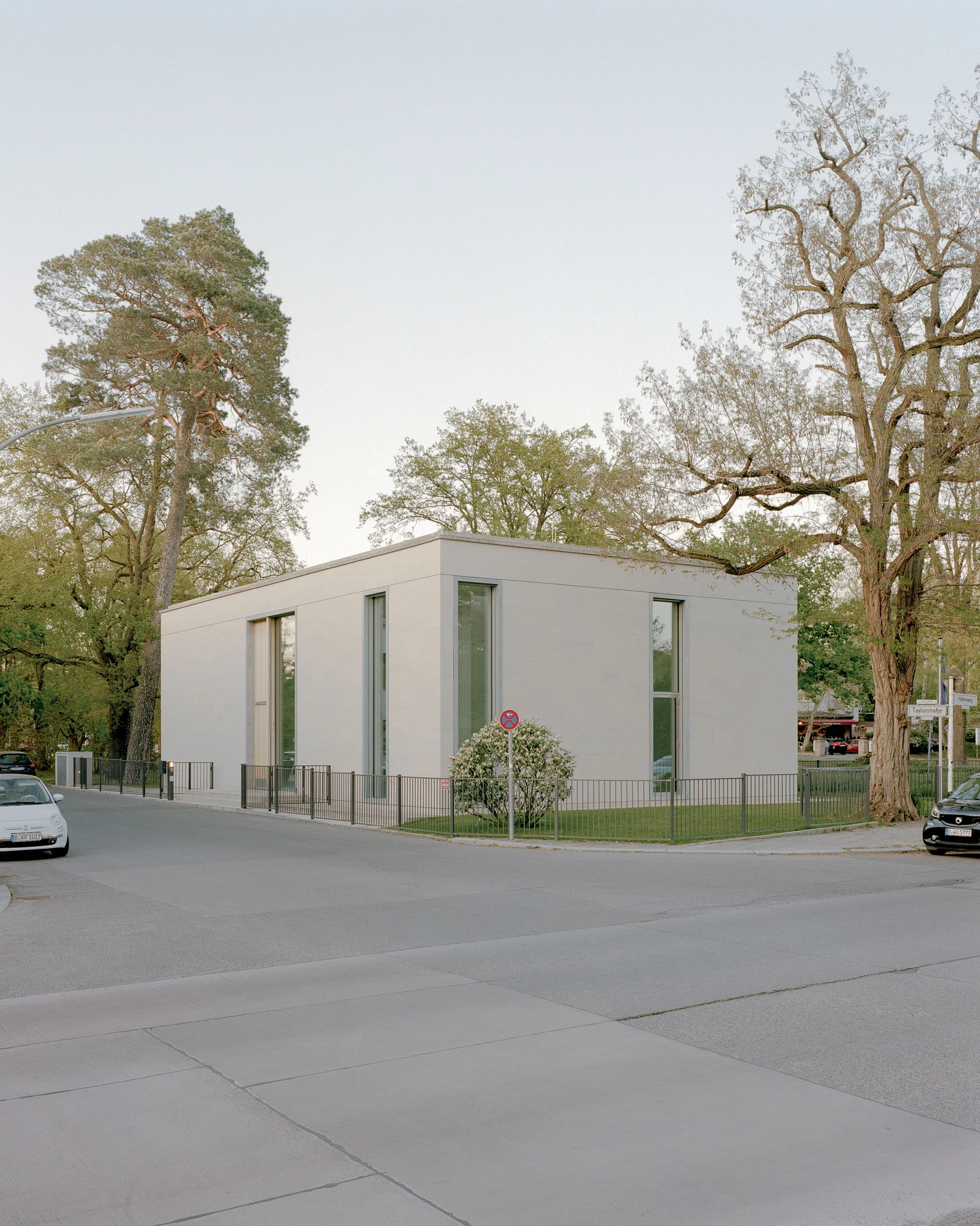 Bastian Gallery by John Pawson, photographed by Julian Holzwarth — Corner view of the Bastian Gallery at dusk: a low sandstone volume with full-height galvanised-steel-framed openings set among bare Berlin park trees, the warm interior glow visible…