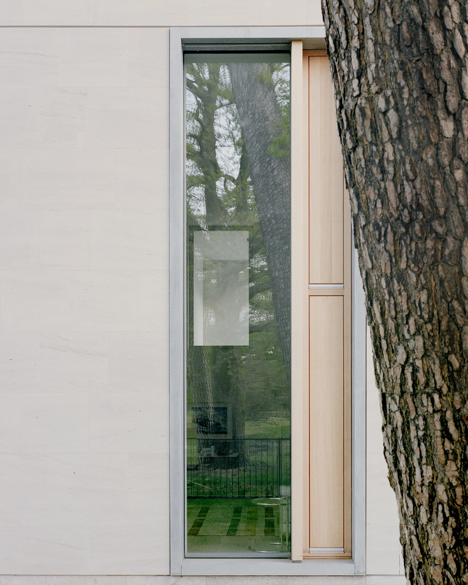 Bastian Gallery by John Pawson, photographed by Julian Holzwarth — Close detail of a full-height opening: galvanised steel frame, vertical oak door panel with visible grain, the reflected image of the park interior showing a mezzanine railing inside.