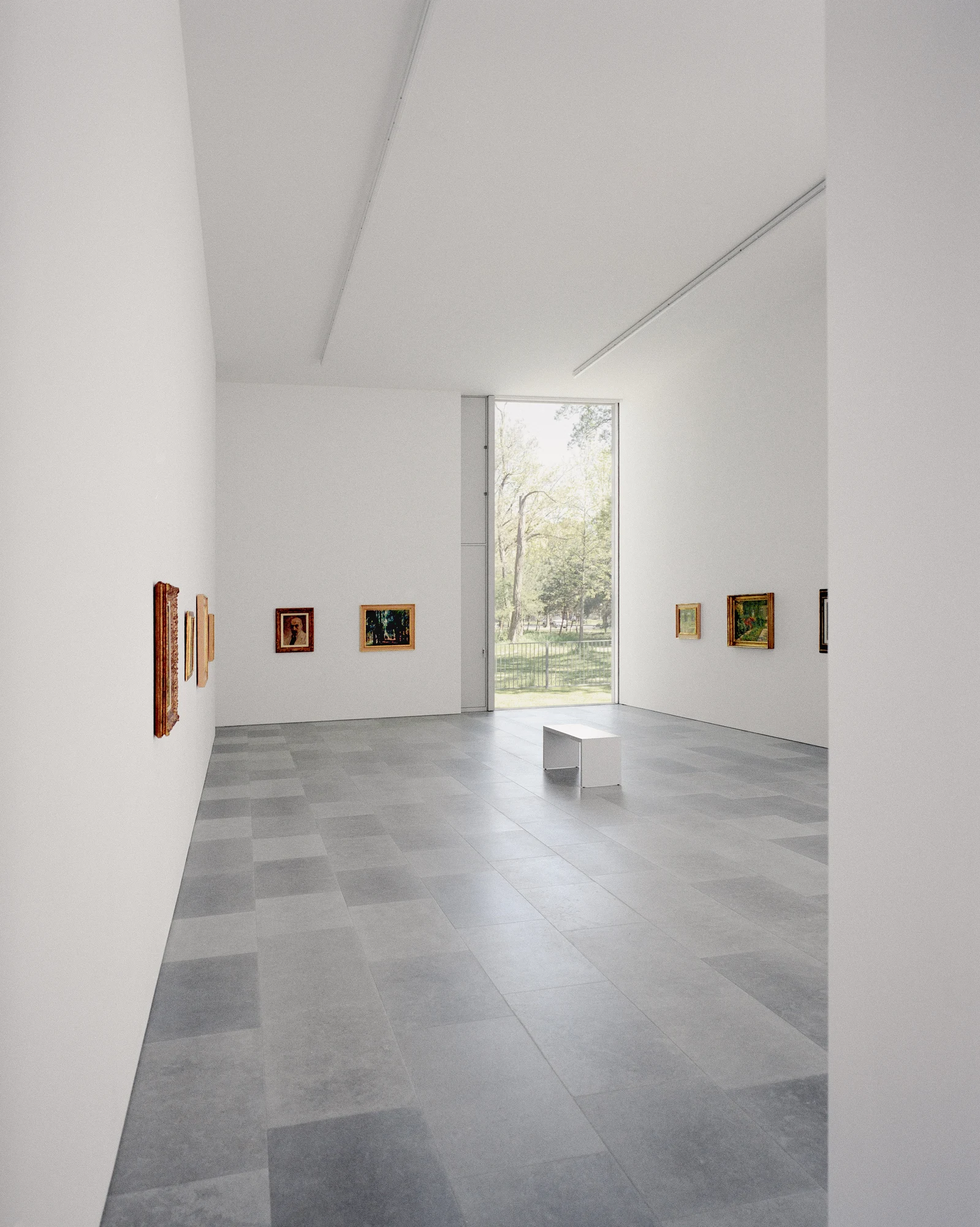 Bastian Gallery by John Pawson, photographed by Julian Holzwarth — Second gallery room with full-height oak-framed glazing opening onto the park: stone tile floor, framed paintings on both walls, a low white bench, the tree canopy directly visible…
