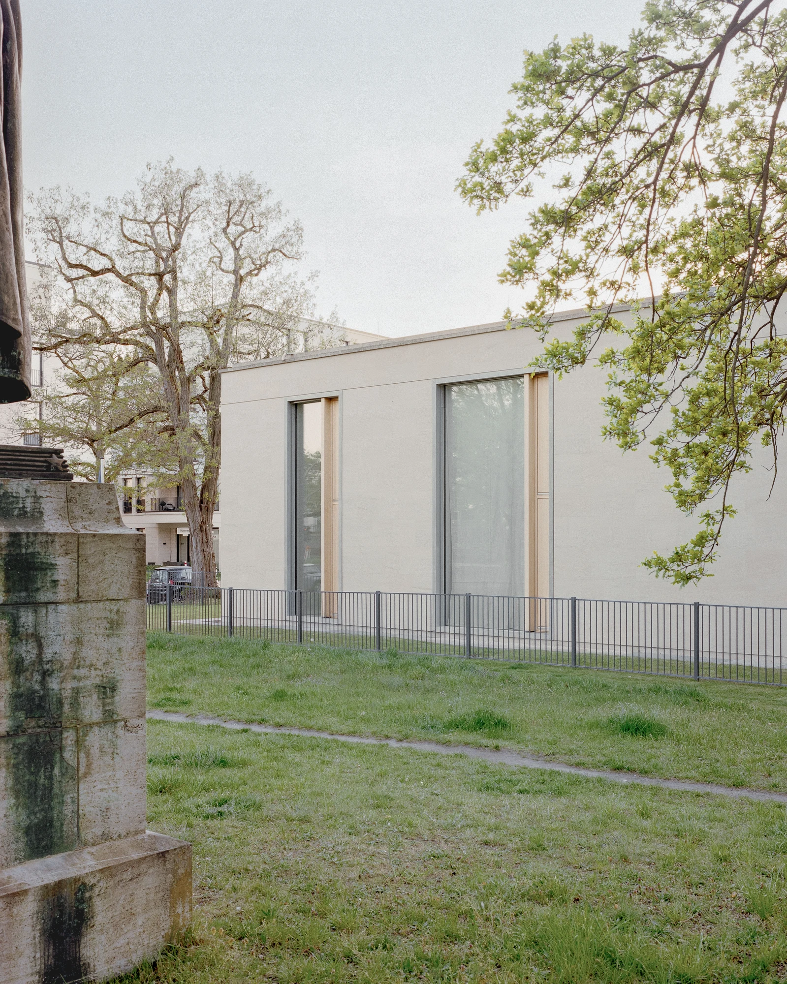 Bastian Gallery by John Pawson, photographed by Julian Holzwarth — Exterior facade in spring light: sandstone cladding, full-height galvanised steel window frames with oak inset panels, a mature tree trunk in the foreground breaking the field of pale…