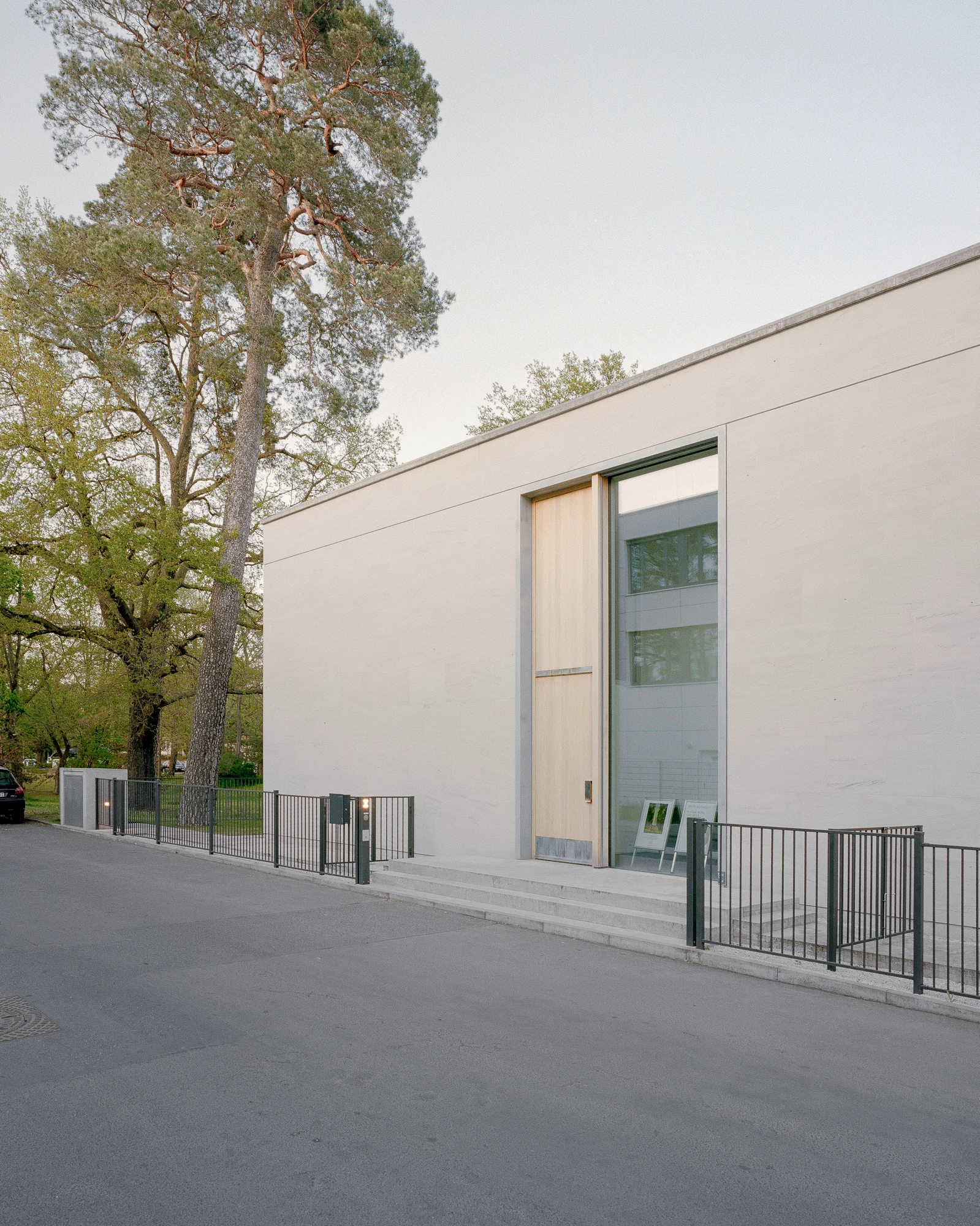 Bastian Gallery by John Pawson, photographed by Julian Holzwarth — Entrance facade: sandstone wall, a full-height oak-framed door panel flanked by glazing, low steps in stone, black steel fence at the boundary, pine tree rising above the parapet.