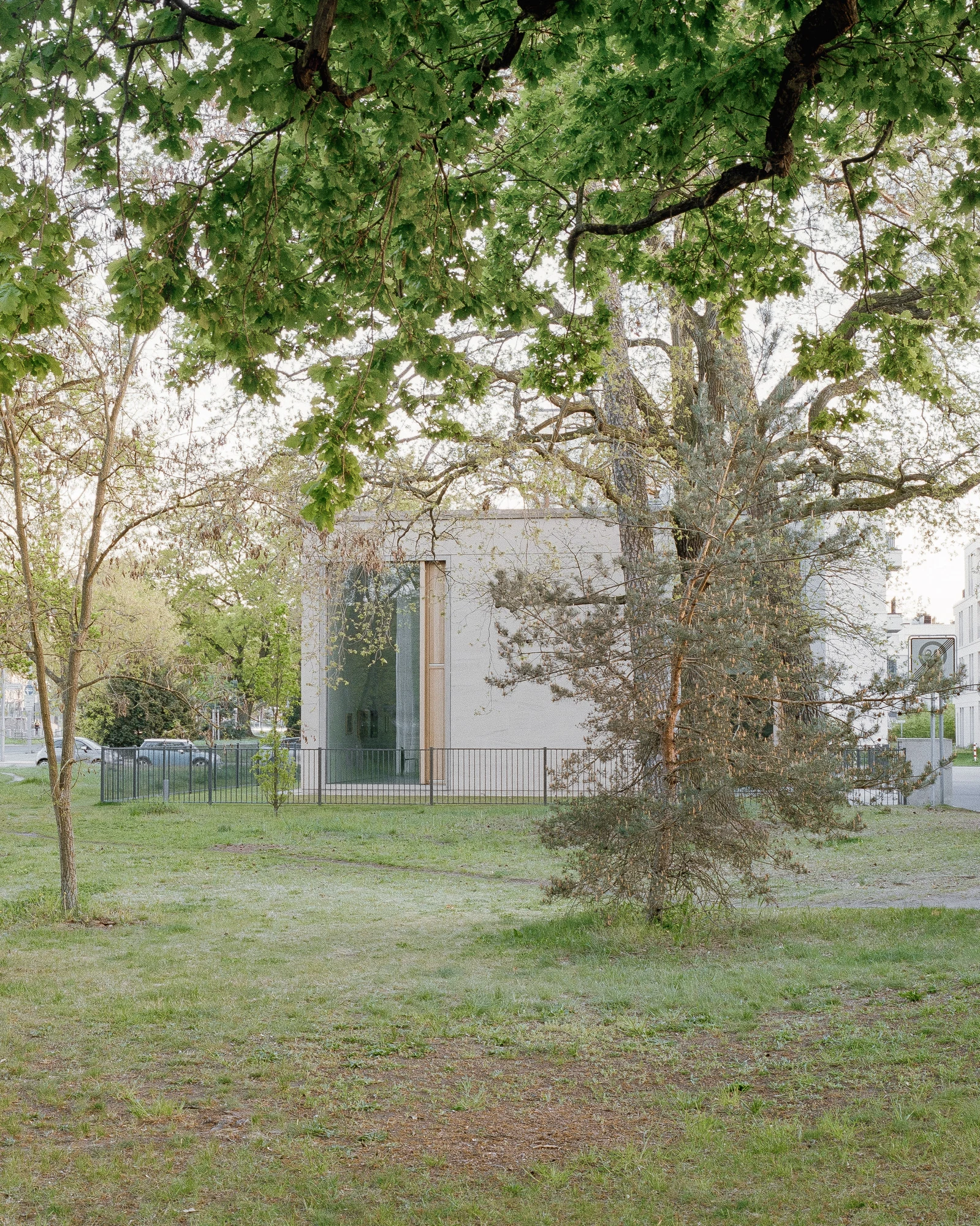 Bastian Gallery by John Pawson, photographed by Julian Holzwarth — Street-facing exterior at dusk: the low sandstone block is framed by a tall pine and a spreading oak, a single full-height entrance opening visible at the building's centre.