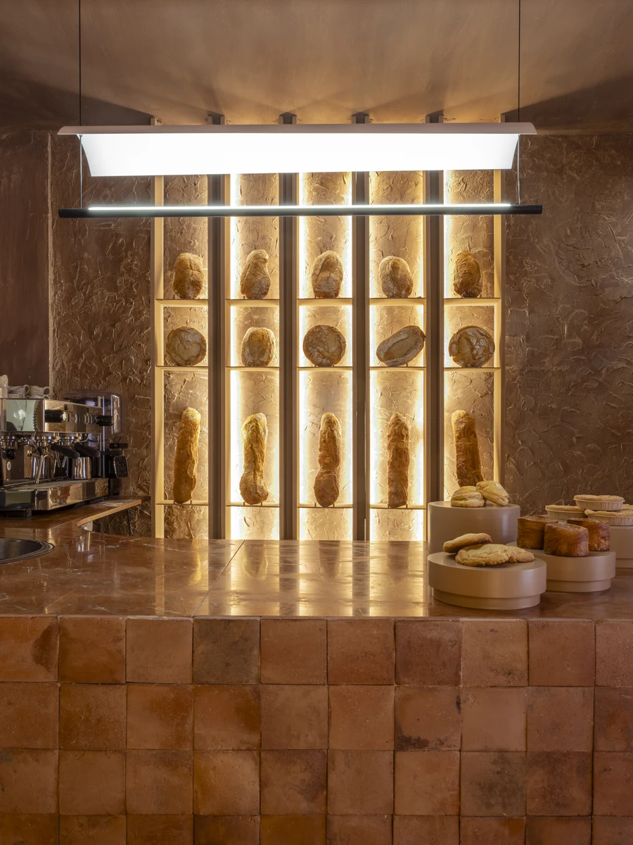 Luchy Bakery Cafe by MOME.ESTUDIO, photographed by Juan Baraja — Close frontal view of the service counter: handmade terracotta tile cladding, a backlit grid shelf holding upright baguettes and round loaves, the amber glow pressing the bread colour into…