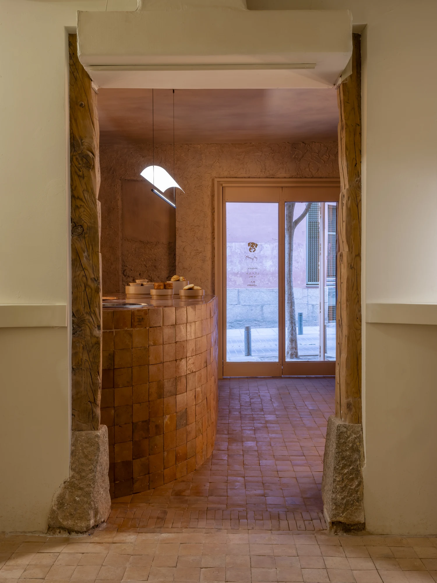 Luchy Bakery Cafe by MOME.ESTUDIO, photographed by Juan Baraja — Room junction showing an original structural timber post set on a raw stone plinth, clay pigment plaster walls, unfired terracotta brick floor in running bond, and a potted plant at the…