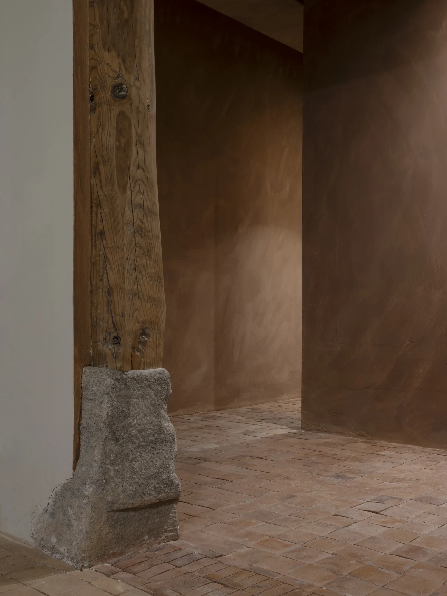 Luchy Bakery Cafe by MOME.ESTUDIO, photographed by Juan Baraja — Rear corner: raw timber post on rough-hewn granite plinth, mid-ochre clay plaster on the walls, terracotta brick pavers on the floor, the junction between two original structural bays left…