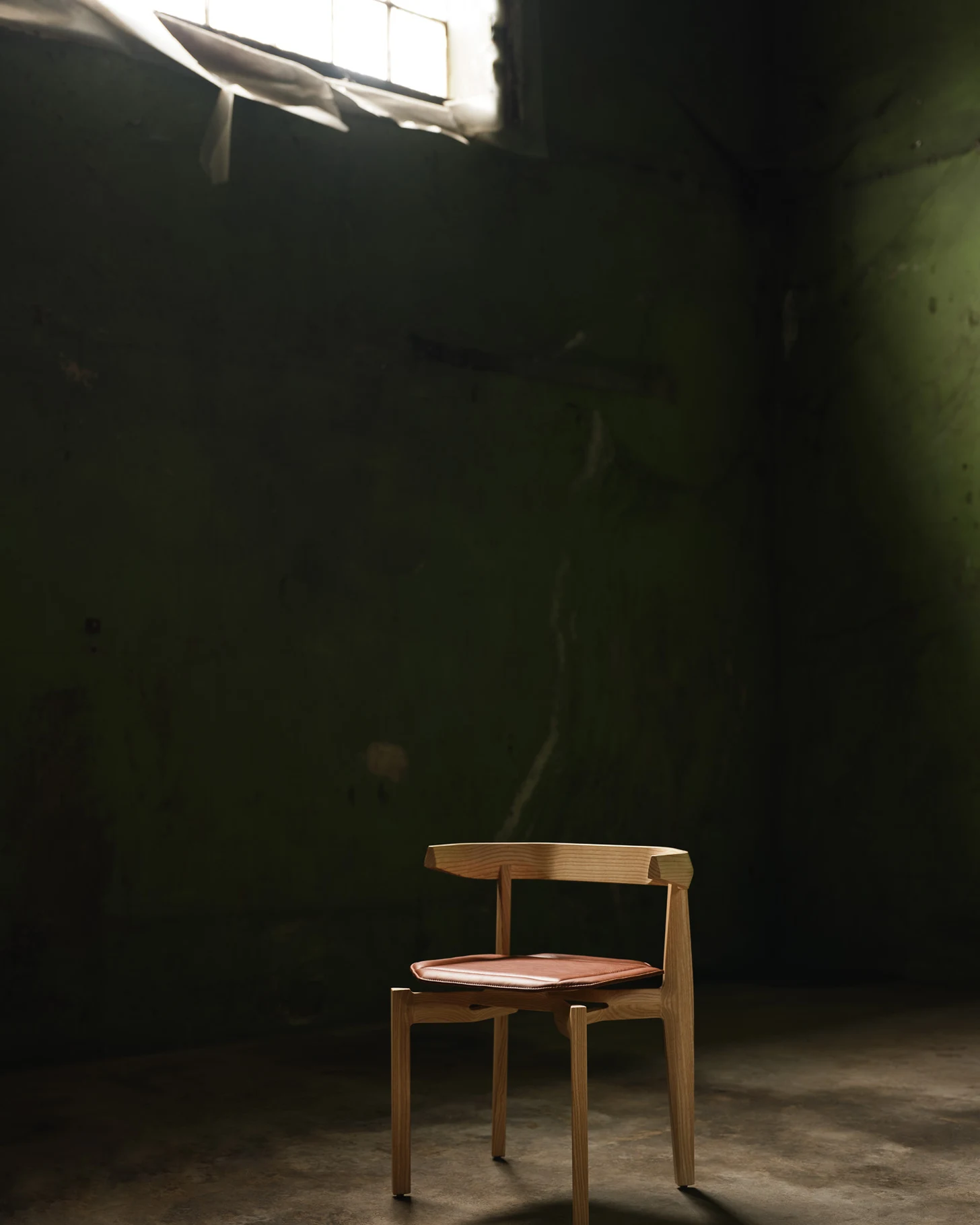 Rasa Collection by Carlos Pereira, photographed by Paulo Carvalho — A single ash dining chair stands on a concrete floor in a derelict warehouse room, pale raking light from a high window revealing the parallel grain lines of the steam-bent backrest and…