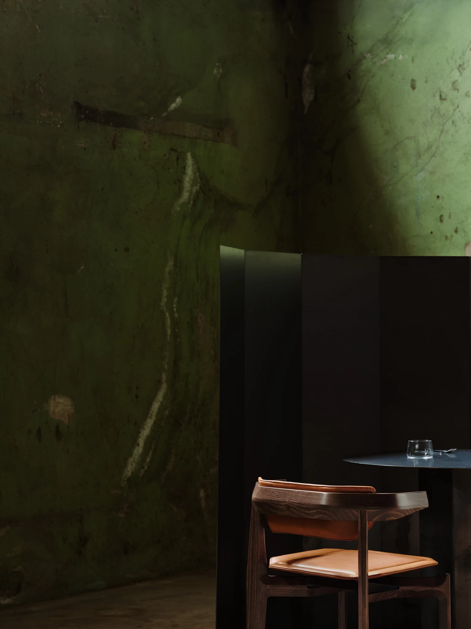 Rasa Collection by Carlos Pereira, photographed by Paulo Carvalho — Close study: a dark stained beech Rasa chair with tan leather seat cushion beside a round black table, the mossy green warehouse wall filling the frame and pressing the warm grain of…