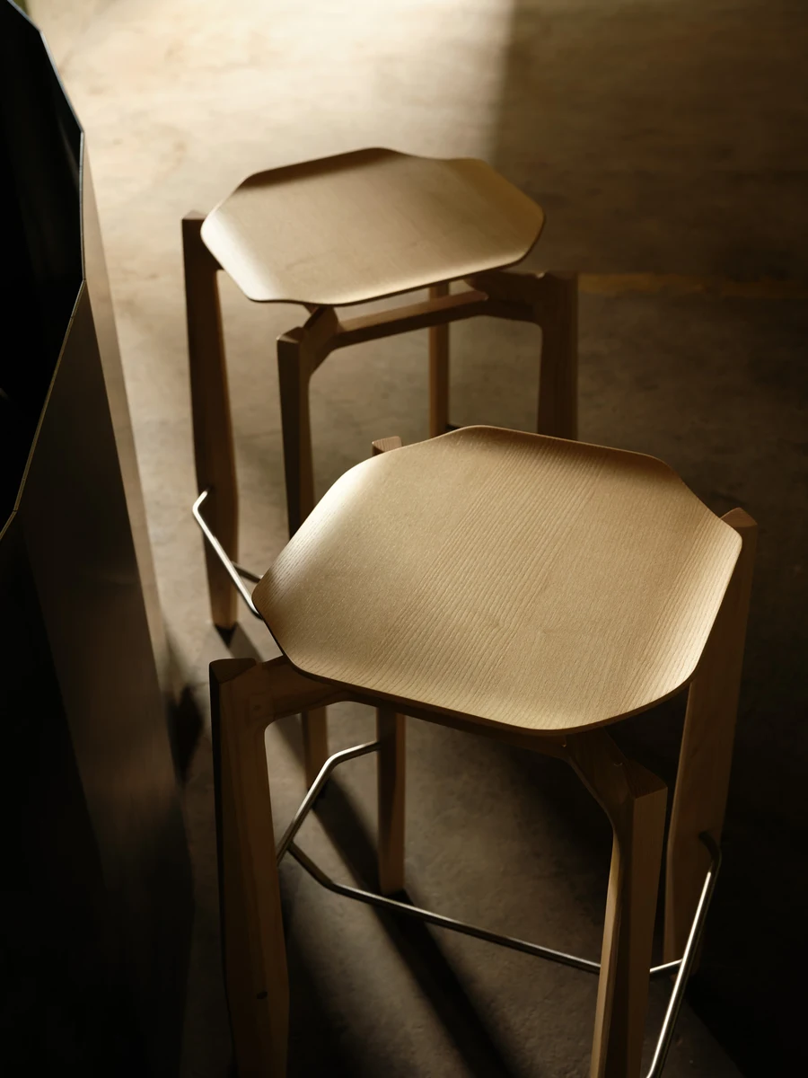 Rasa Collection by Carlos Pereira, photographed by Paulo Carvalho — Two natural ash bar stools photographed from above: the octagonal seat shells show the parallel lines of steam-bent grain under warm directional light, the bent steel footrest ring…