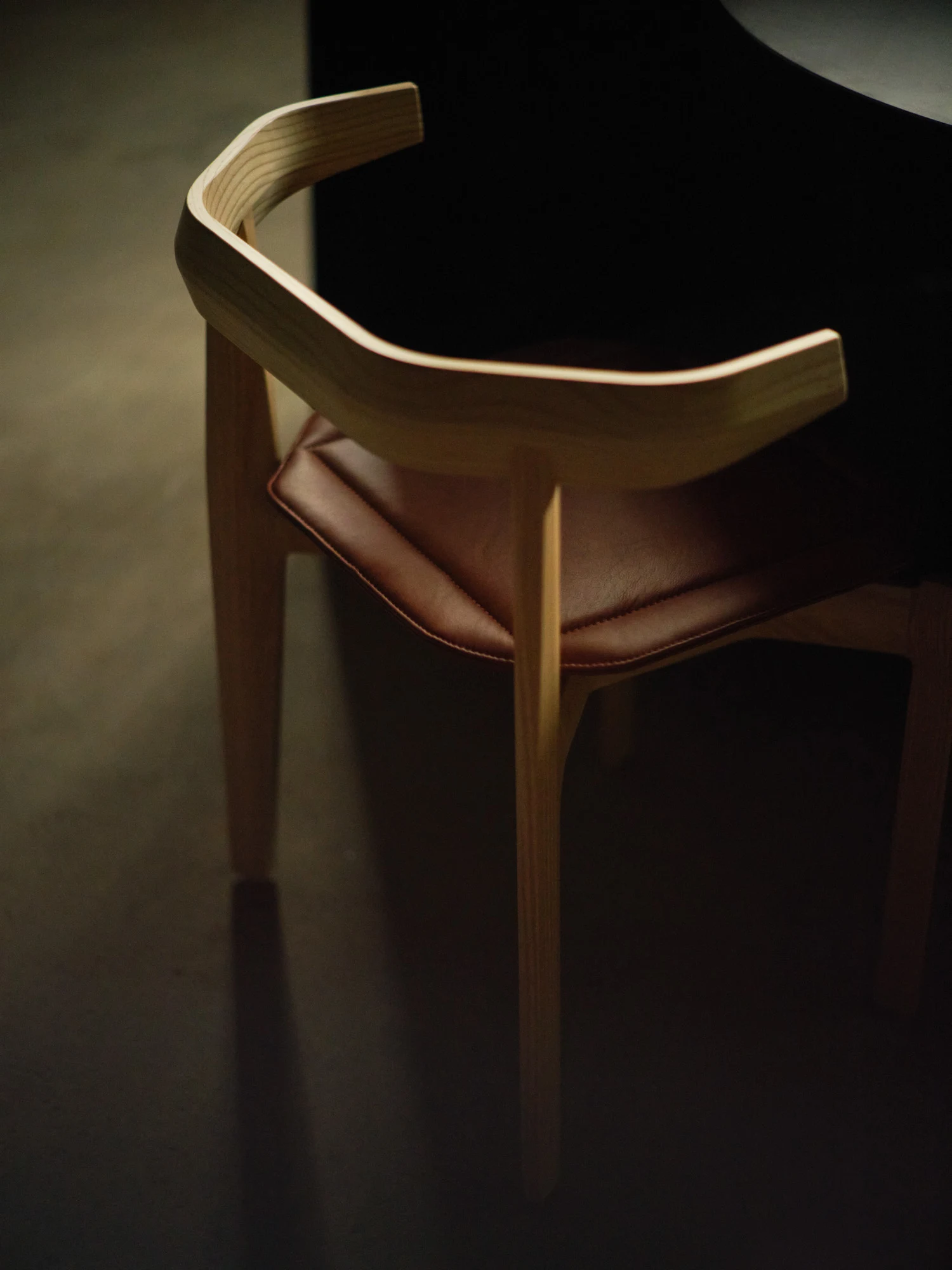 Rasa Collection by Carlos Pereira, photographed by Paulo Carvalho — Natural ash Rasa chair on concrete floor against a dark warehouse wall: the full profile of the piece, four legs, mortised seat shell, single-plank backrest, visible in three-quarter…