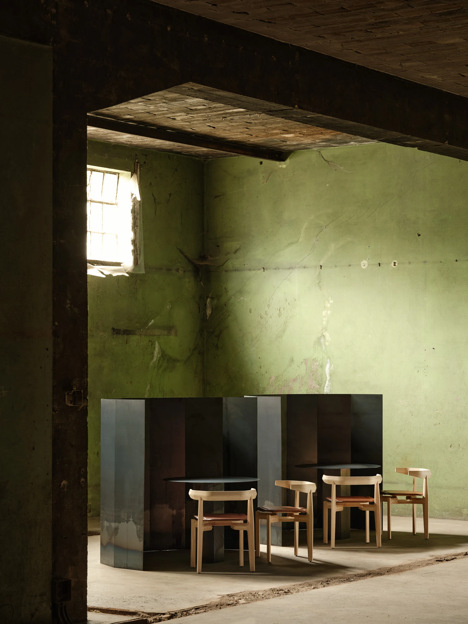 Rasa Collection by Carlos Pereira, photographed by Paulo Carvalho — Corner of the warehouse with dark steel partitions and Rasa dining chairs with orange upholstered cushions arranged around round black tables, peeling green plaster wall and brick…