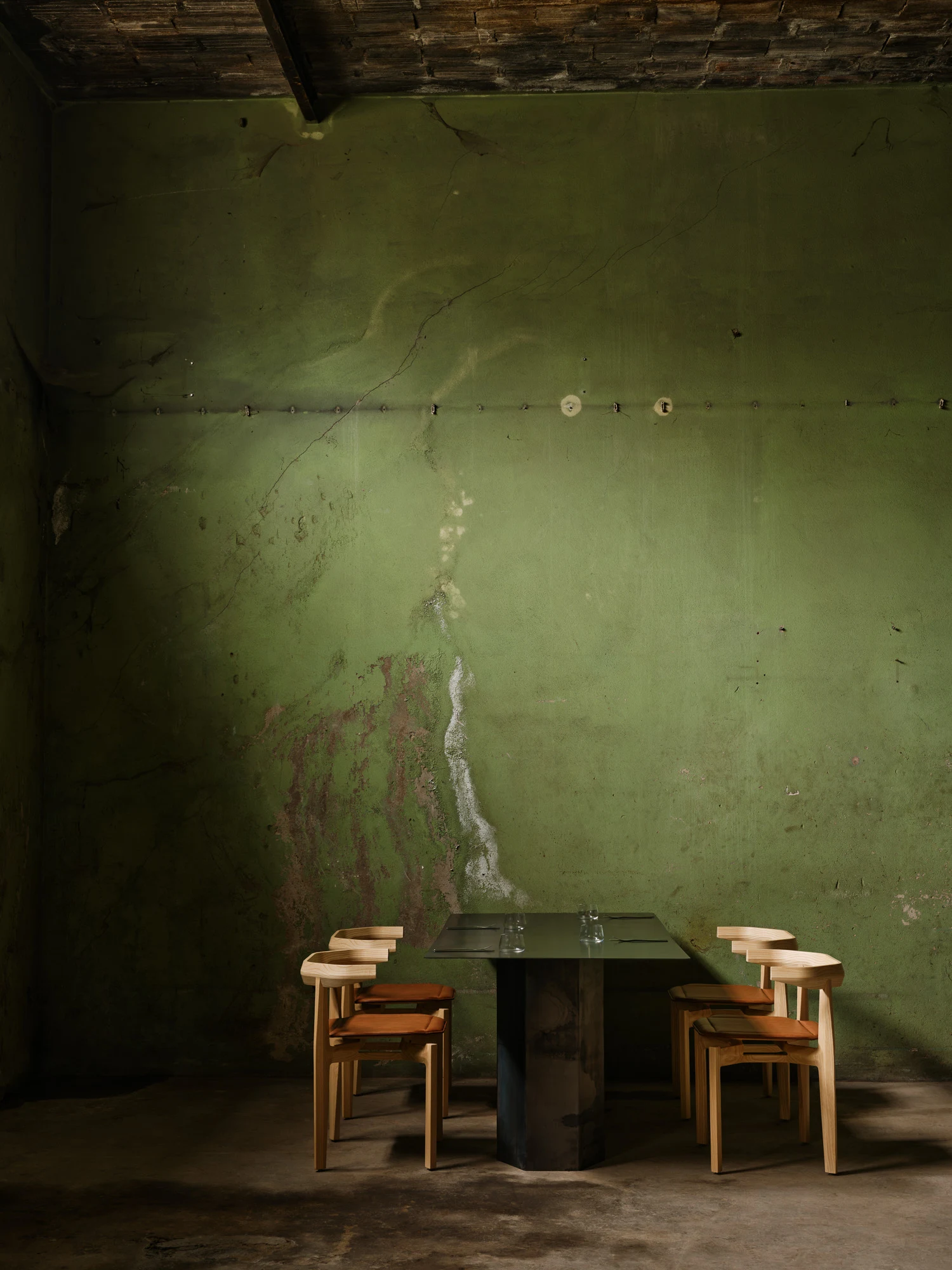 Rasa Collection by Carlos Pereira, photographed by Paulo Carvalho — Four Rasa dining chairs with orange cushions around a rectangular black table centred against the green warehouse wall, the scene isolated and frontal, the concrete floor showing its age.