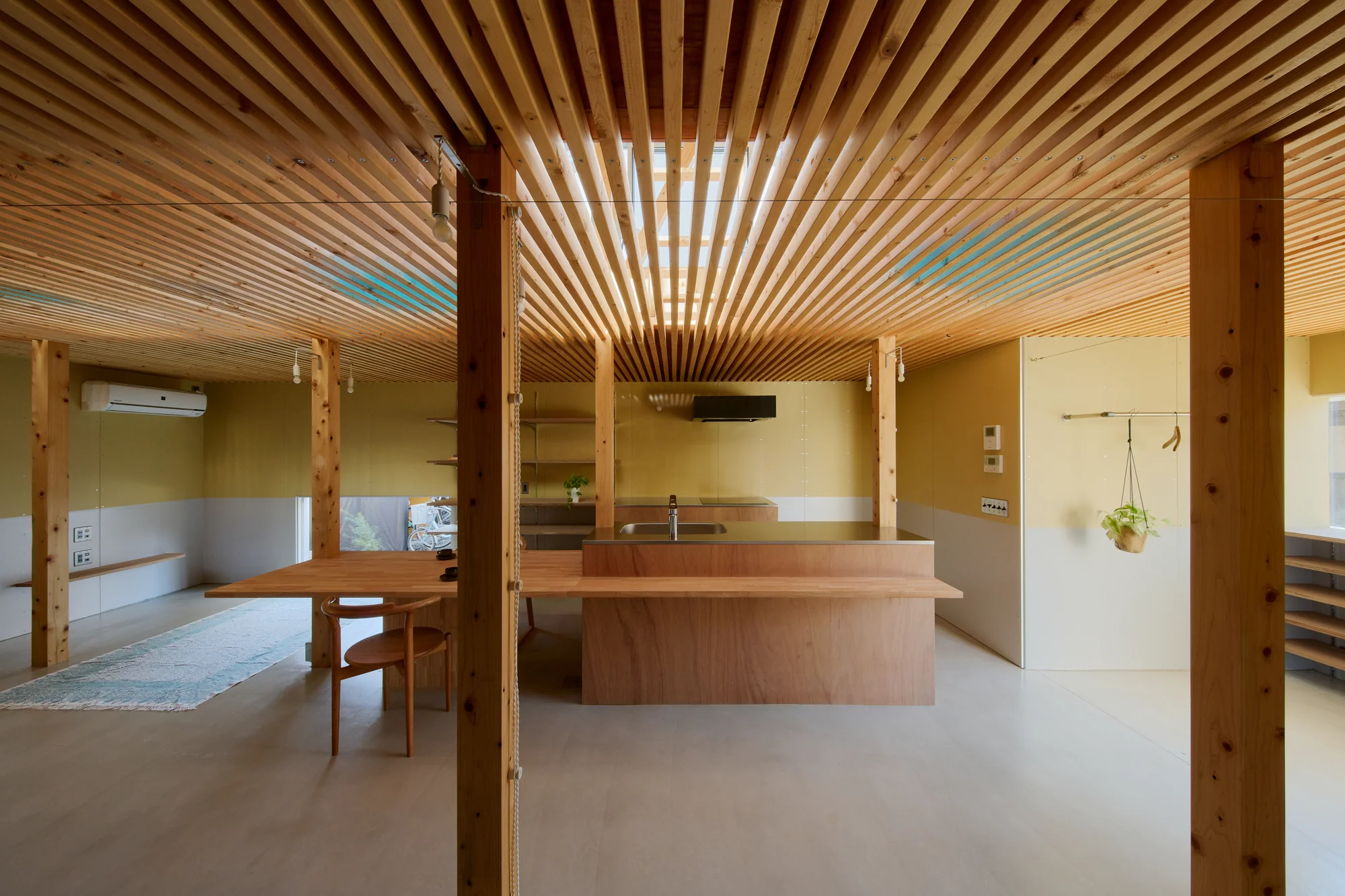 House TN by 1-1 Architects, photographed by Takashi Uemura — Kitchen and living zone under the continuous timber louver ceiling, plywood island centered between timber columns, walls painted in two-tone olive and grey.