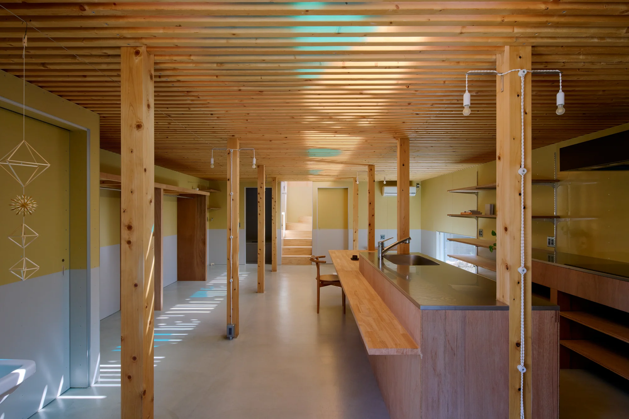 House TN by 1-1 Architects, photographed by Takashi Uemura — Ground floor interior with 2x4 timber louver ceiling filtering light from above, timber columns, plywood kitchen counter, olive walls meeting glossy earthen floor.