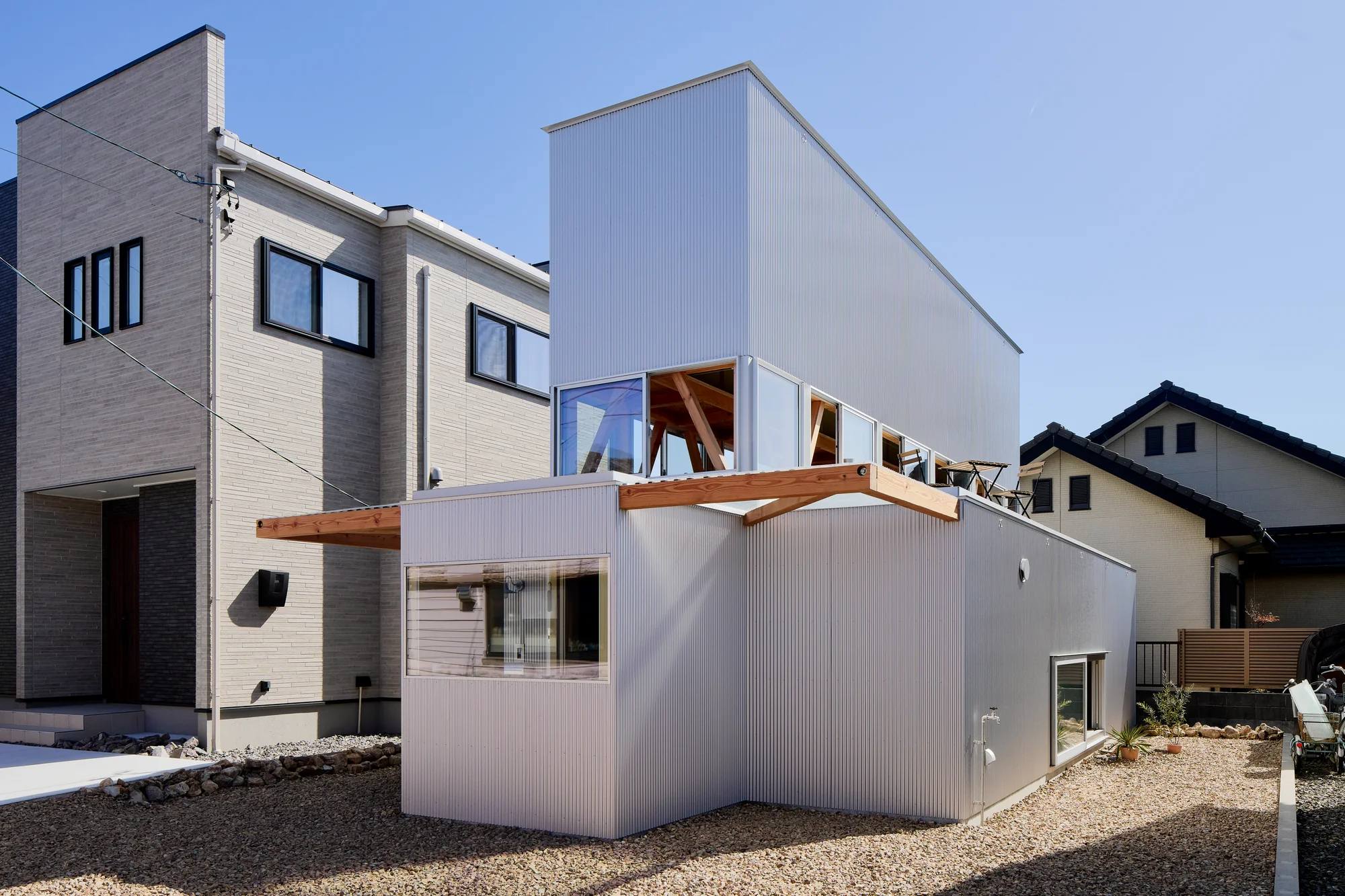 House TN by 1-1 Architects, photographed by Takashi Uemura — Corner view shows the stepped massing: ground-floor box in corrugated metal, setback second floor with glazed openings, gravel yard below.