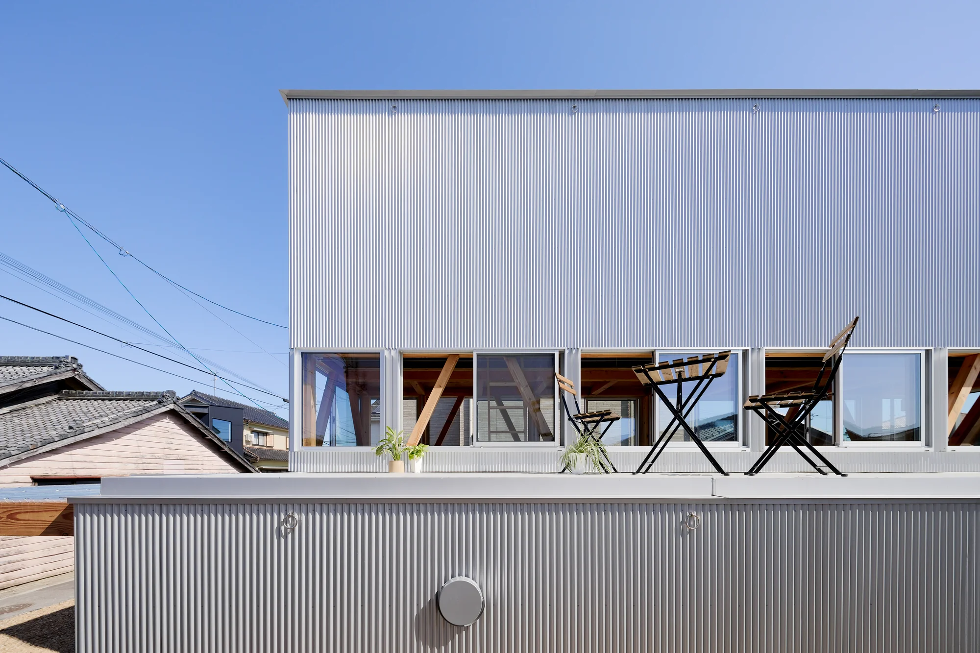 House TN by 1-1 Architects, photographed by Takashi Uemura — The second-floor terrace holds bistro furniture, continuous window band revealing the timber structure inside, neighboring tile roofs visible beyond.