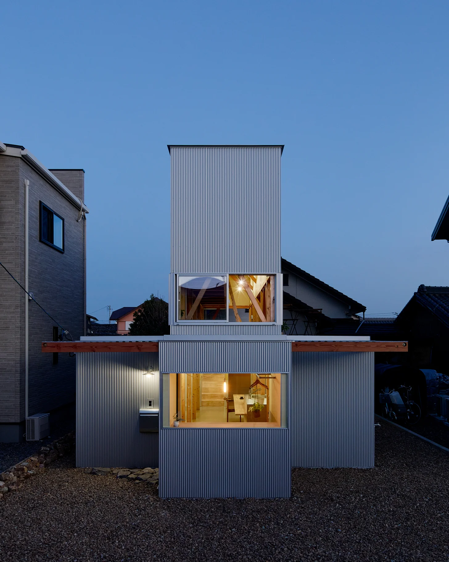 House TN by 1-1 Architects, photographed by Takashi Uemura — Corrugated metal volumes stack at dusk, warm timber glow visible through ground and upper-floor windows against the blue hour sky.