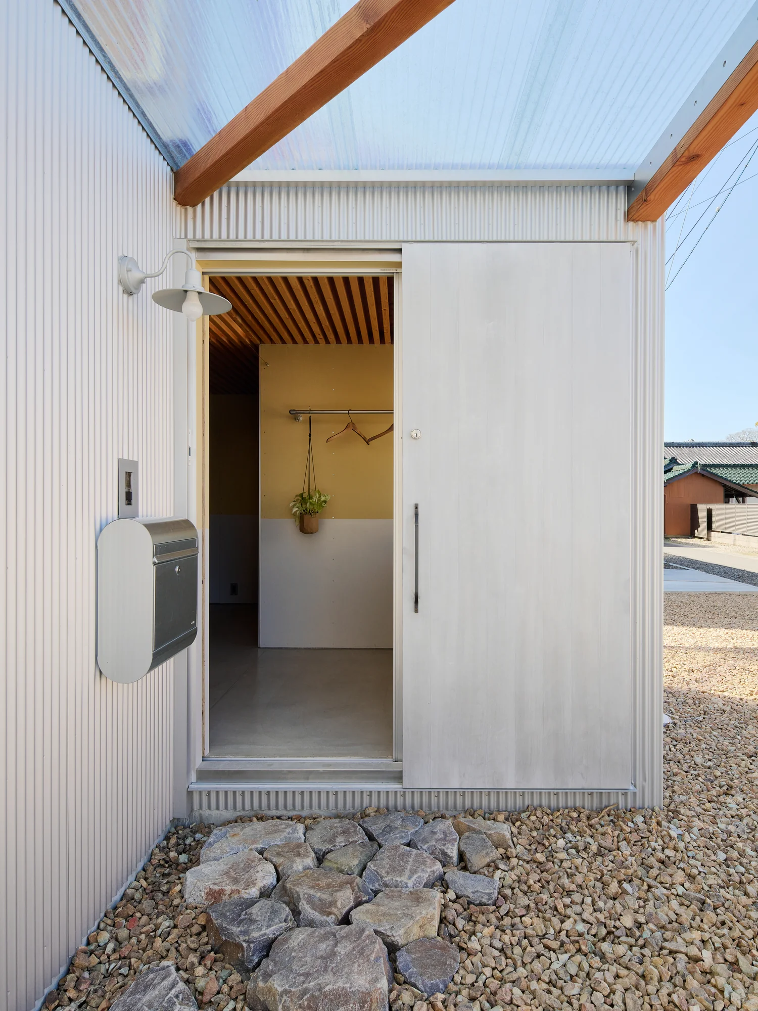 House TN by 1-1 Architects, photographed by Takashi Uemura — Entry sequence: corrugated metal walls frame a timber-louver canopy, rough stone path leading to the sliding door.