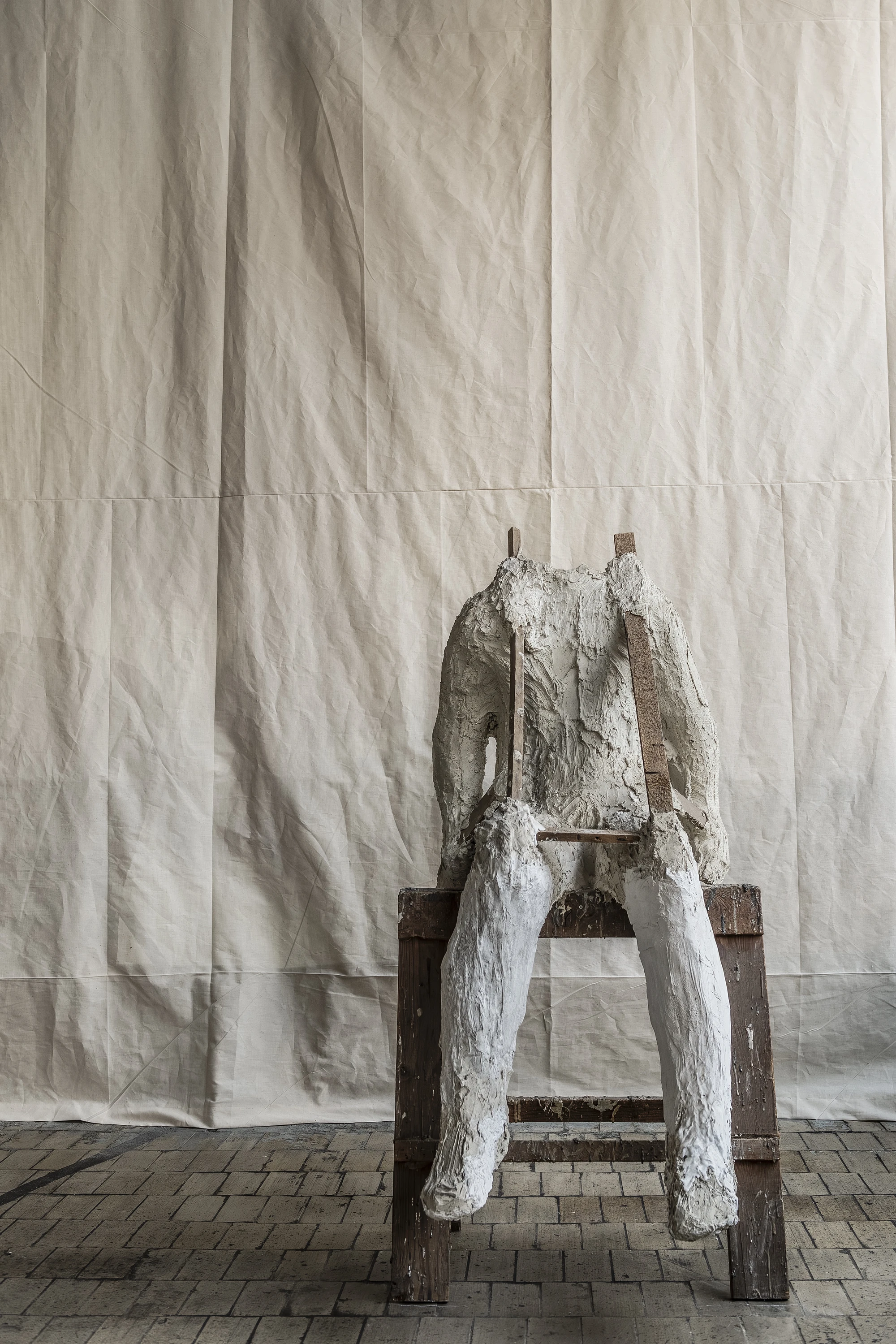 Magdalena Abakanowicz / Wanda Czełkowska by Abakanowicz Foundation, photographed by Norbert Piwowarczyk — a single plaster figure from the Tłum series, seated on a low timber stool against a creased white sheet.