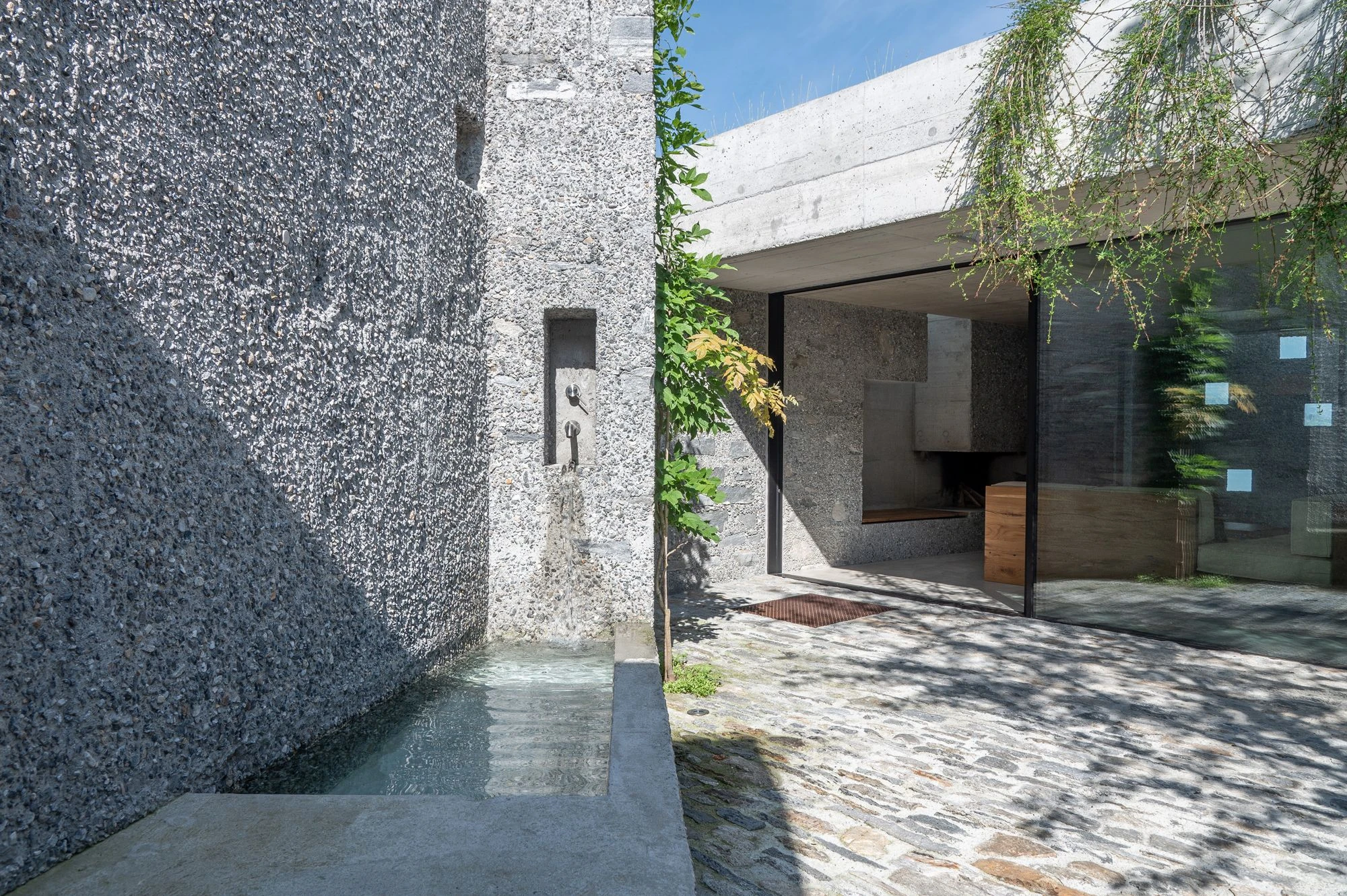 New House in San Nazzaro by Wespi de Meuron Romeo, photographed by Giacomo Albo — the entrance courtyard with a fountain trough and oak-framed glazing onto the living space.