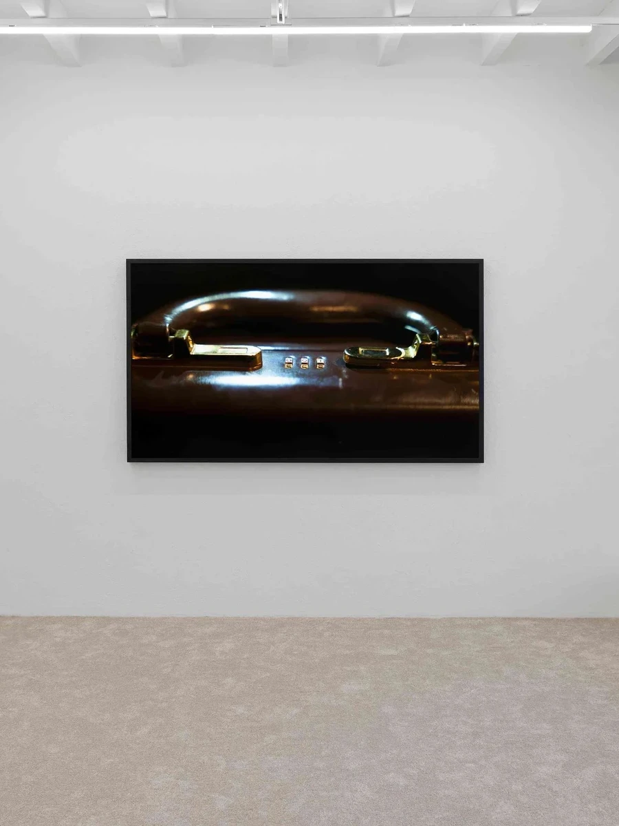 Installation view at Blue Velvet Projects, Zürich, showing a widescreen framed still by Flavio Karrer of a brass-locked briefcase handle hung on a pale wall above beige carpet.