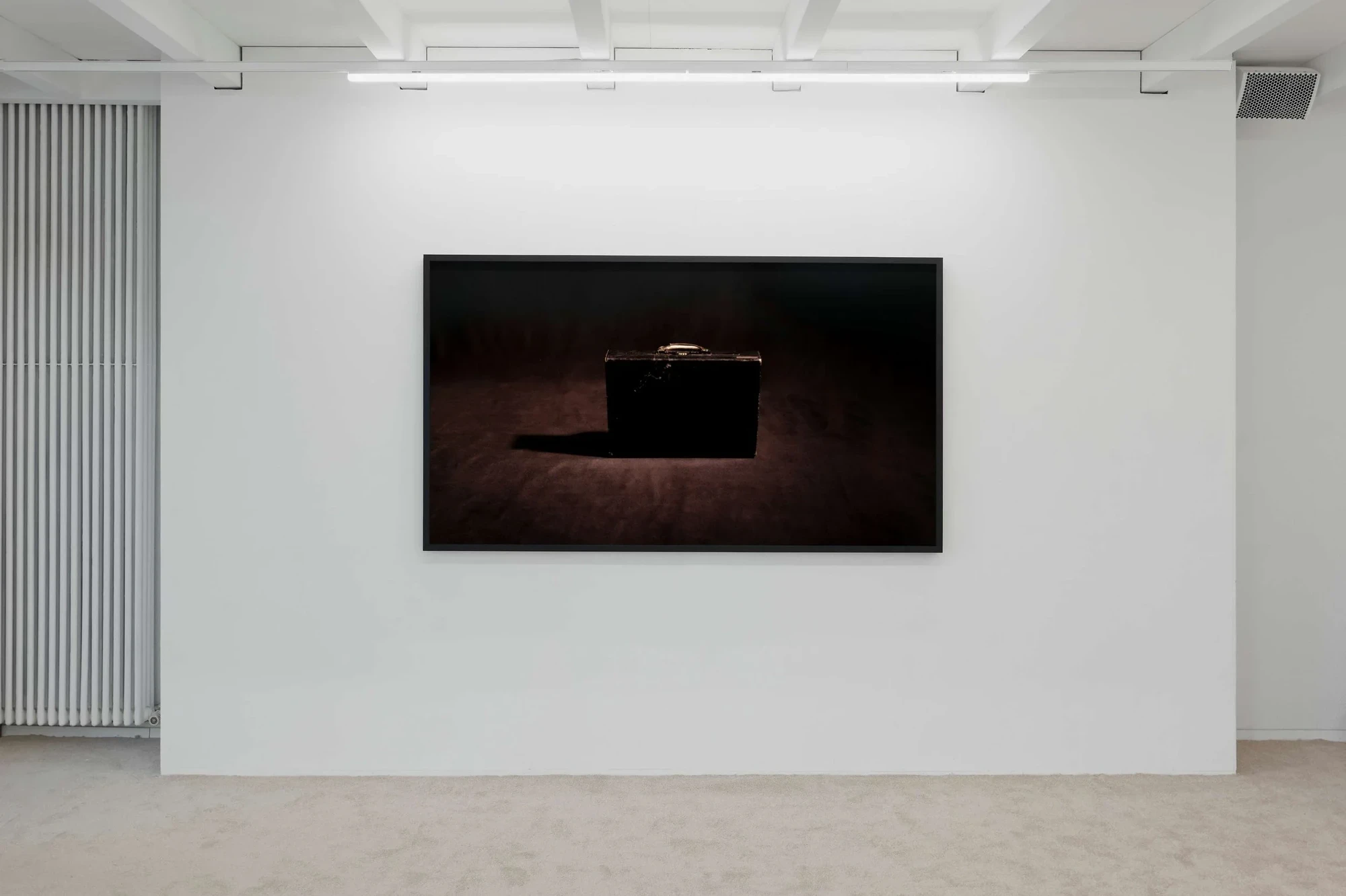 A large framed photograph of a single dark attaché briefcase on a maroon ground, hung on a white wall beside a vertical louvred radiator.