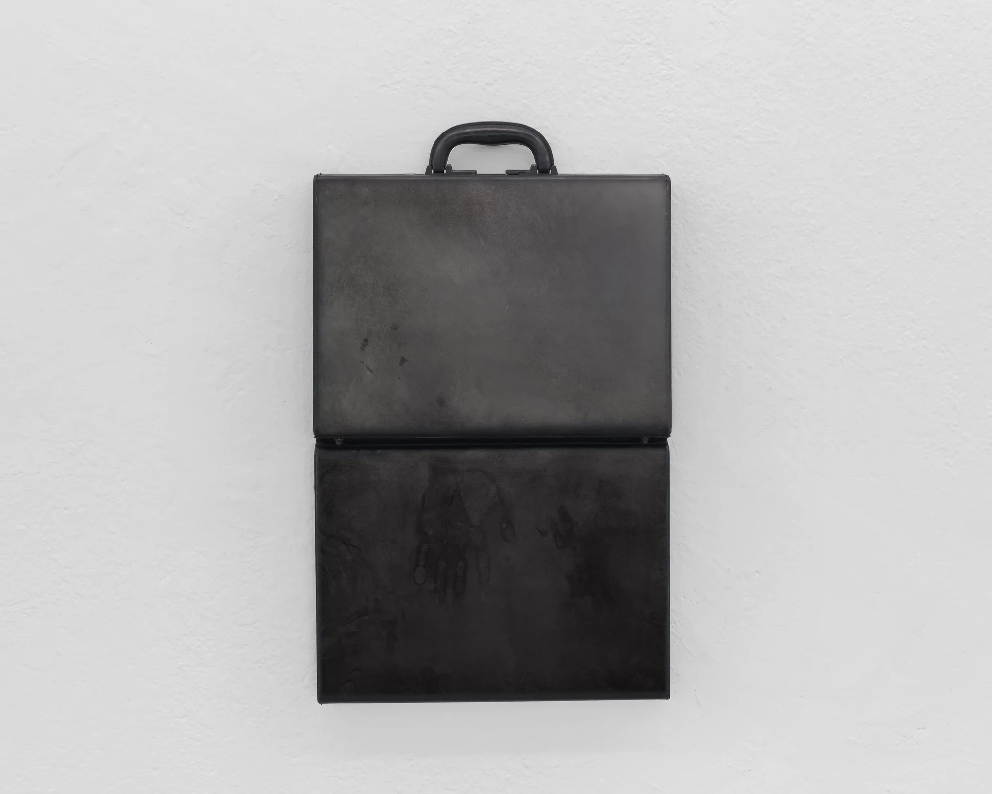 A matte black attaché case mounted to the wall with its lid folded forward, faint handprint-like marks ghosting the lower interior surface.