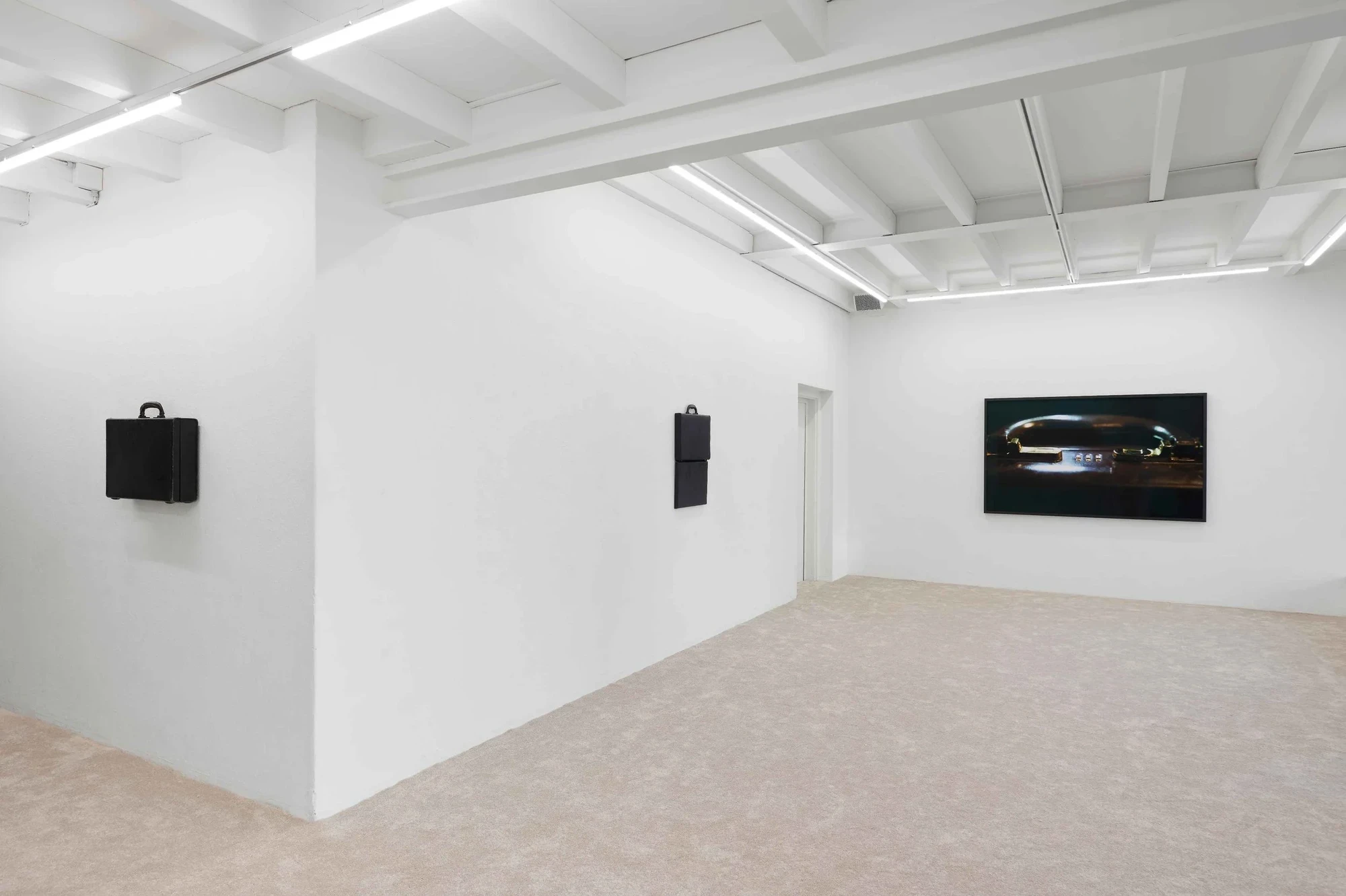 Gallery view at Blue Velvet Projects in Zürich with two black wall-mounted briefcase sculptures and a framed photograph by Flavio Karrer on the far wall.