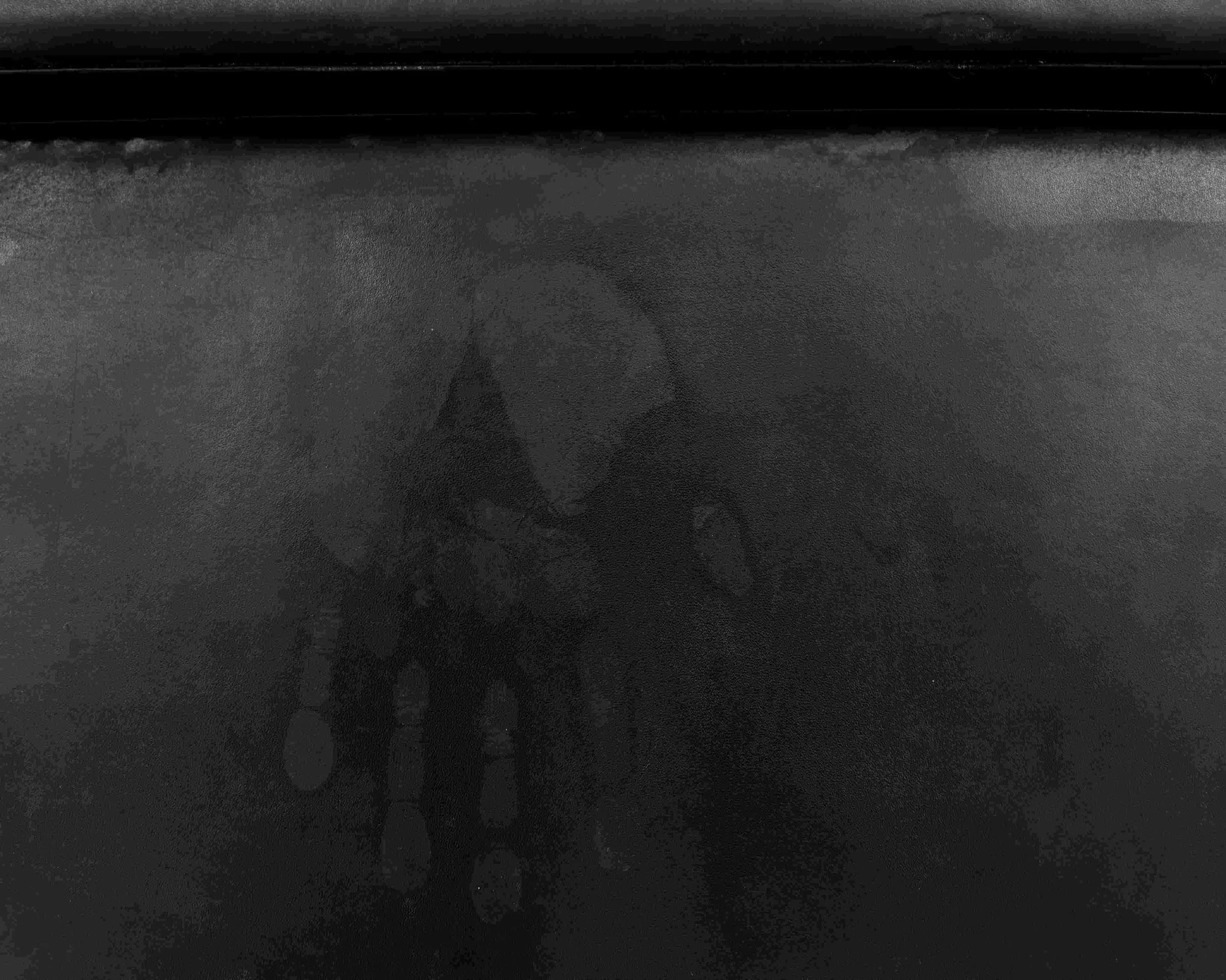 Detail of a black briefcase surface mottled with milky drips and faint hand-shaped traces, the residue catching low light.
