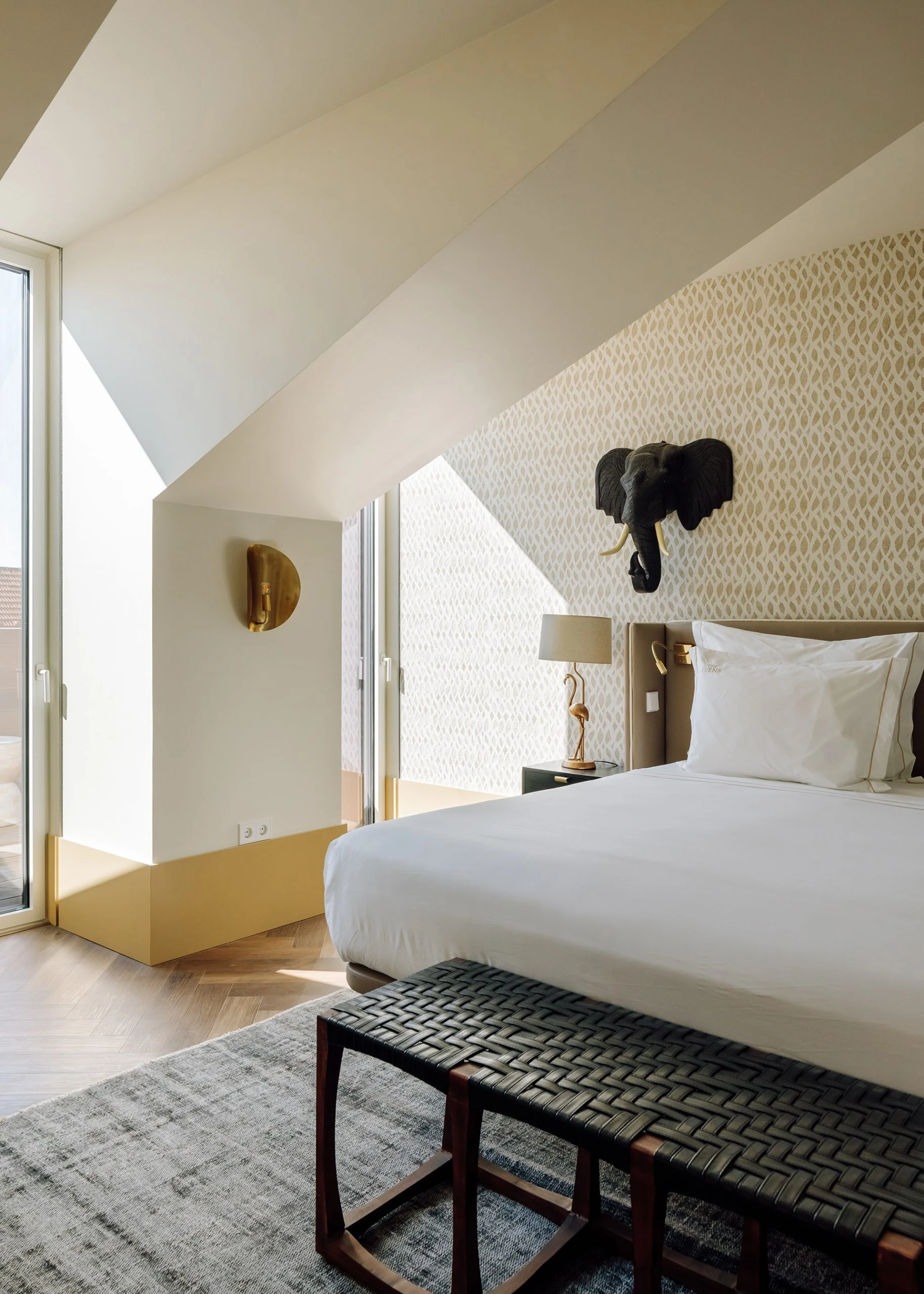 The Ivens by Lázaro Rosa-Violan, photographed by Francisco Nogueira — mansard suite with mounted ceramic elephant, small-pattern wallpaper and plaited leather bench.