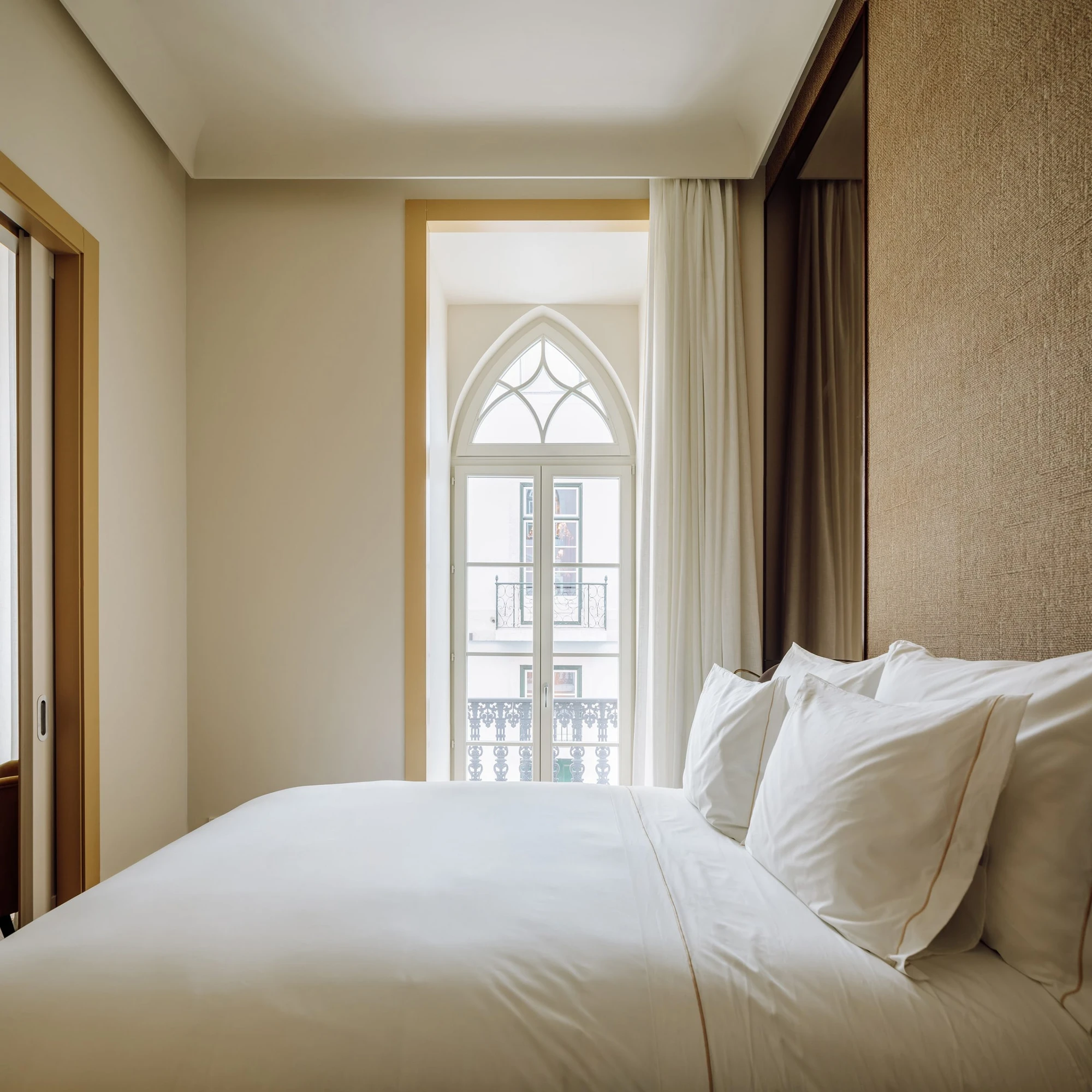 The Ivens by Lázaro Rosa-Violan, photographed by Francisco Nogueira — guest room with pale oak window surround framing a neo-Gothic arched window and a jute headboard panel.