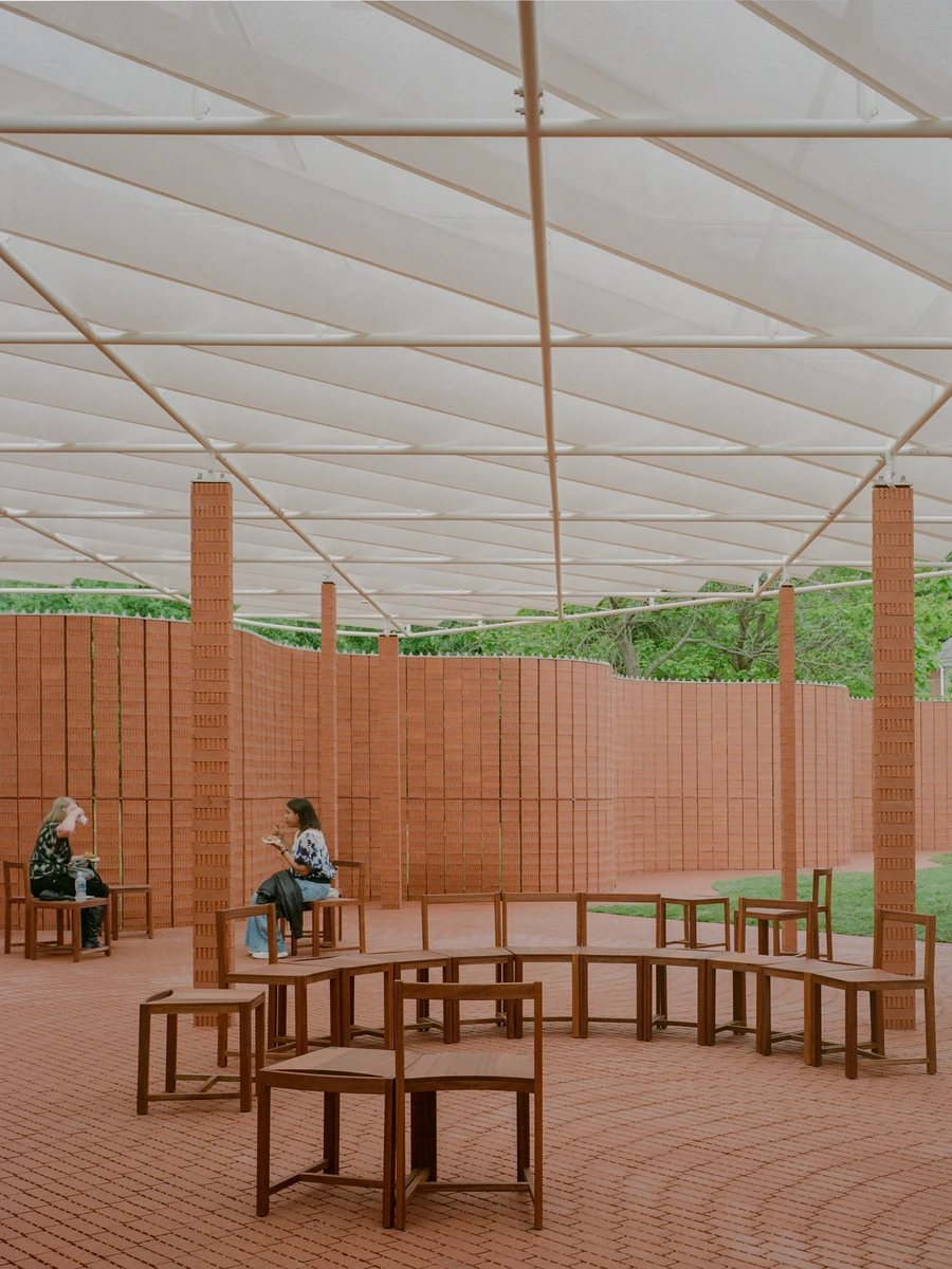 Serpentine Pavilion 2026 by LANZA atelier, photographed by Lorenzo Zandri — visitors rest on walnut-toned timber benches inside the brick enclosure under the diffused light of the fabric canopy.