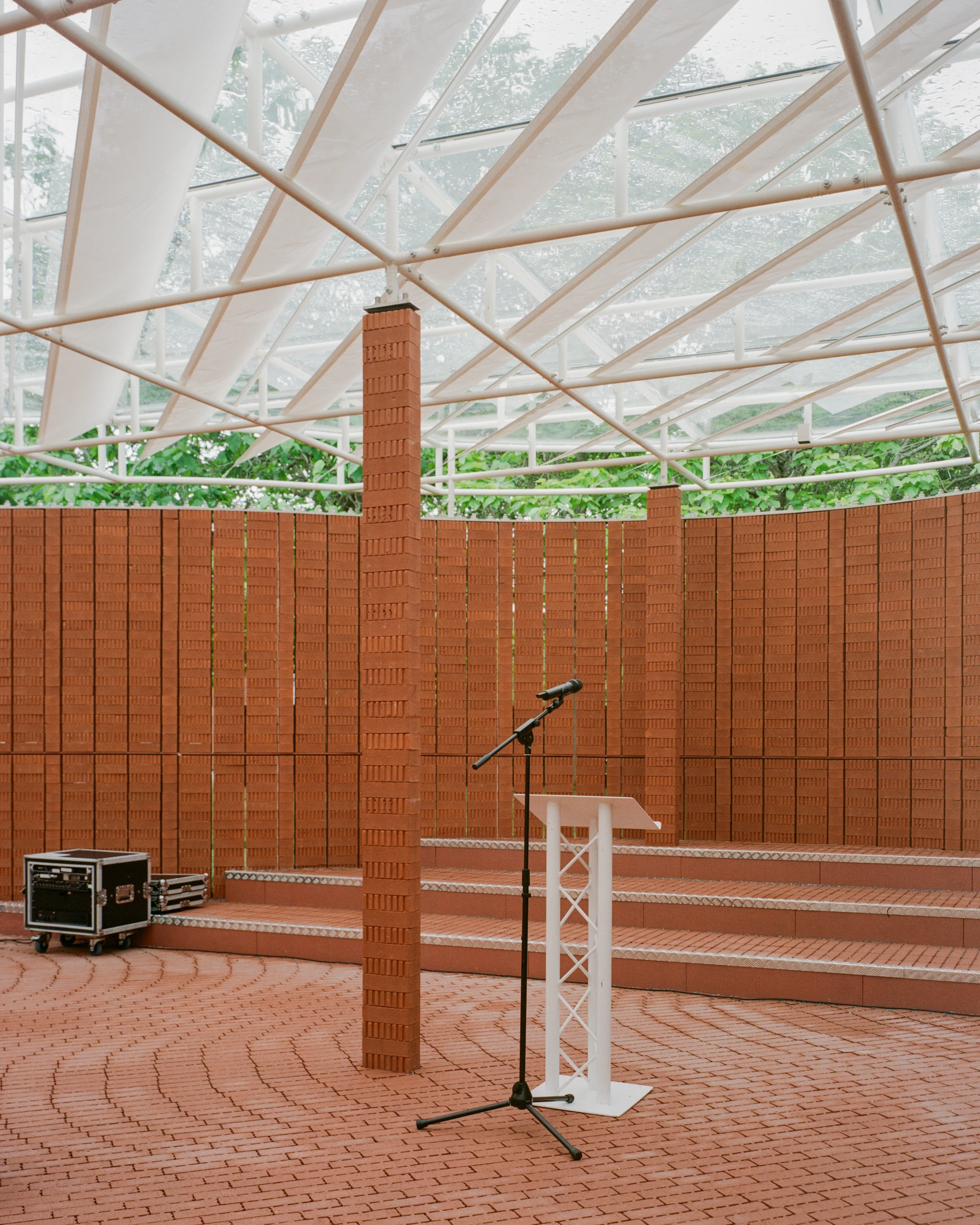 Serpentine Pavilion 2026 by LANZA atelier, photographed by Lorenzo Zandri — a microphone and lectern set for a reading, the brick steps and column lit from above by the translucent fabric.