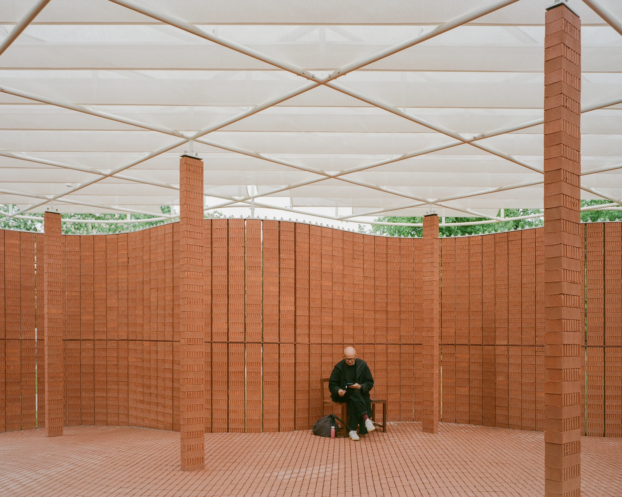 Serpentine Pavilion 2026 by LANZA atelier, photographed by Lorenzo Zandri — a visitor seated alone between two brick columns, the diffused fabric roof crossing overhead in a grid of steel rods.