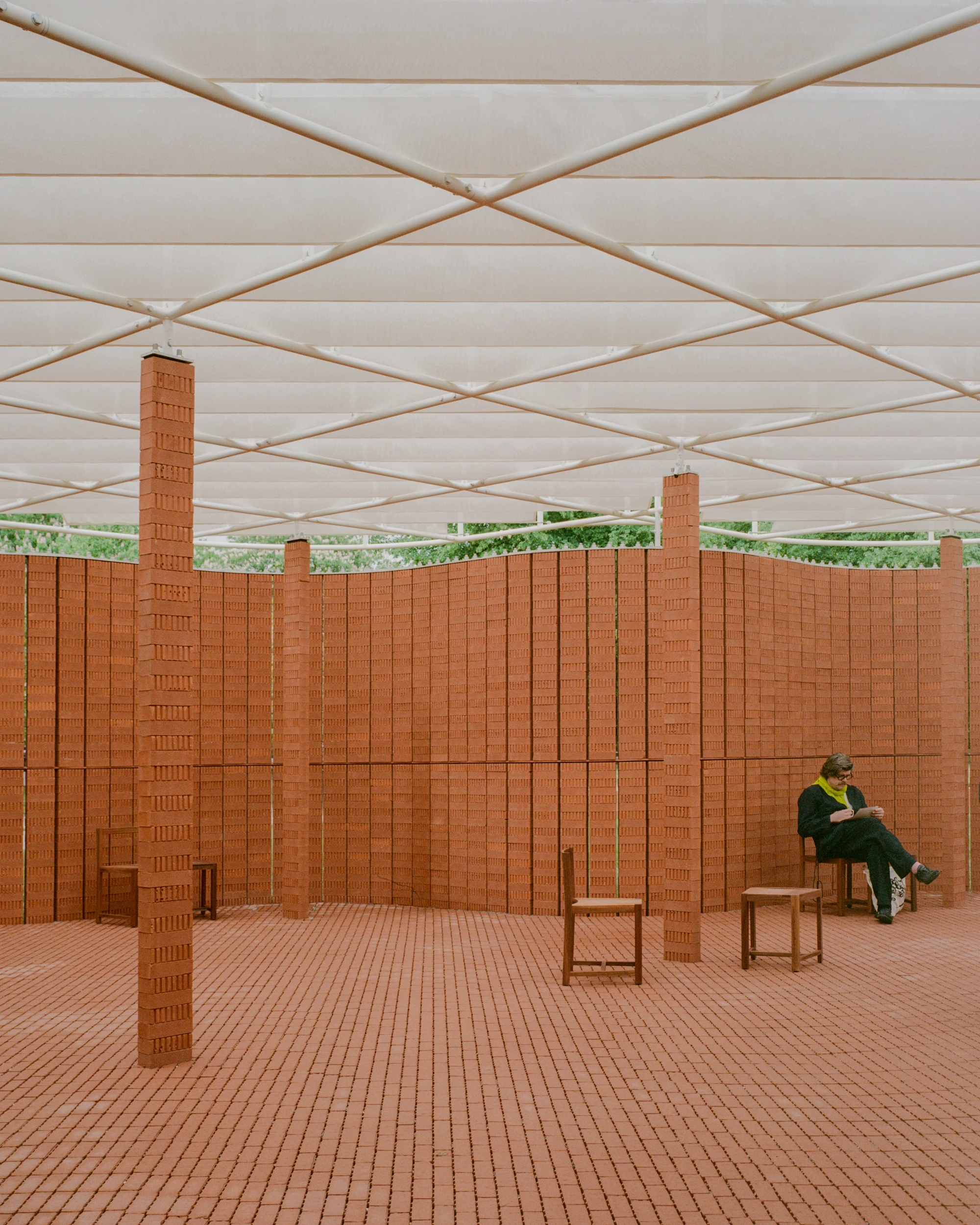 Serpentine Pavilion 2026 by LANZA atelier, photographed by Lorenzo Zandri — interior view with brick columns rising to the white space frame and a visitor reading on a timber chair against the perforated wall.