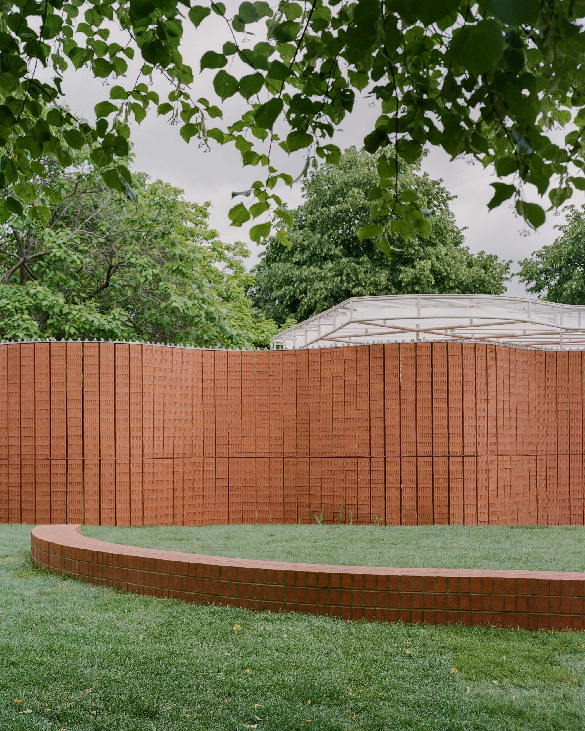 Serpentine Pavilion 2026 by LANZA atelier, photographed by Lorenzo Zandri — the pavilion seen through hanging leaves, with the low brick bench tracing a wide arc on the grass.