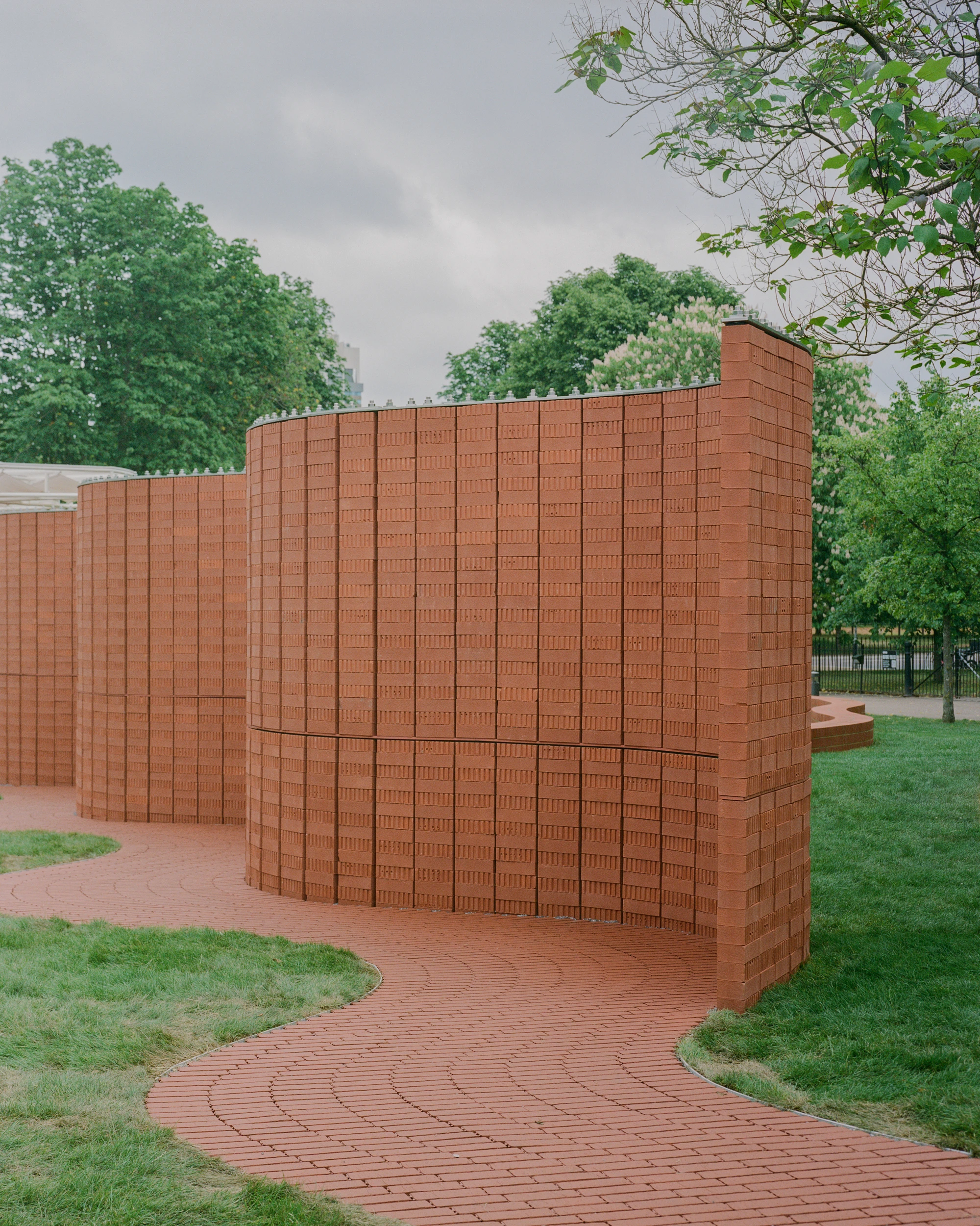 Serpentine Pavilion 2026 by LANZA atelier, photographed by Lorenzo Zandri — the south wall bends in a tight curve where the brick path enters the pavilion, with the brick columns rising clear of the enclosure.