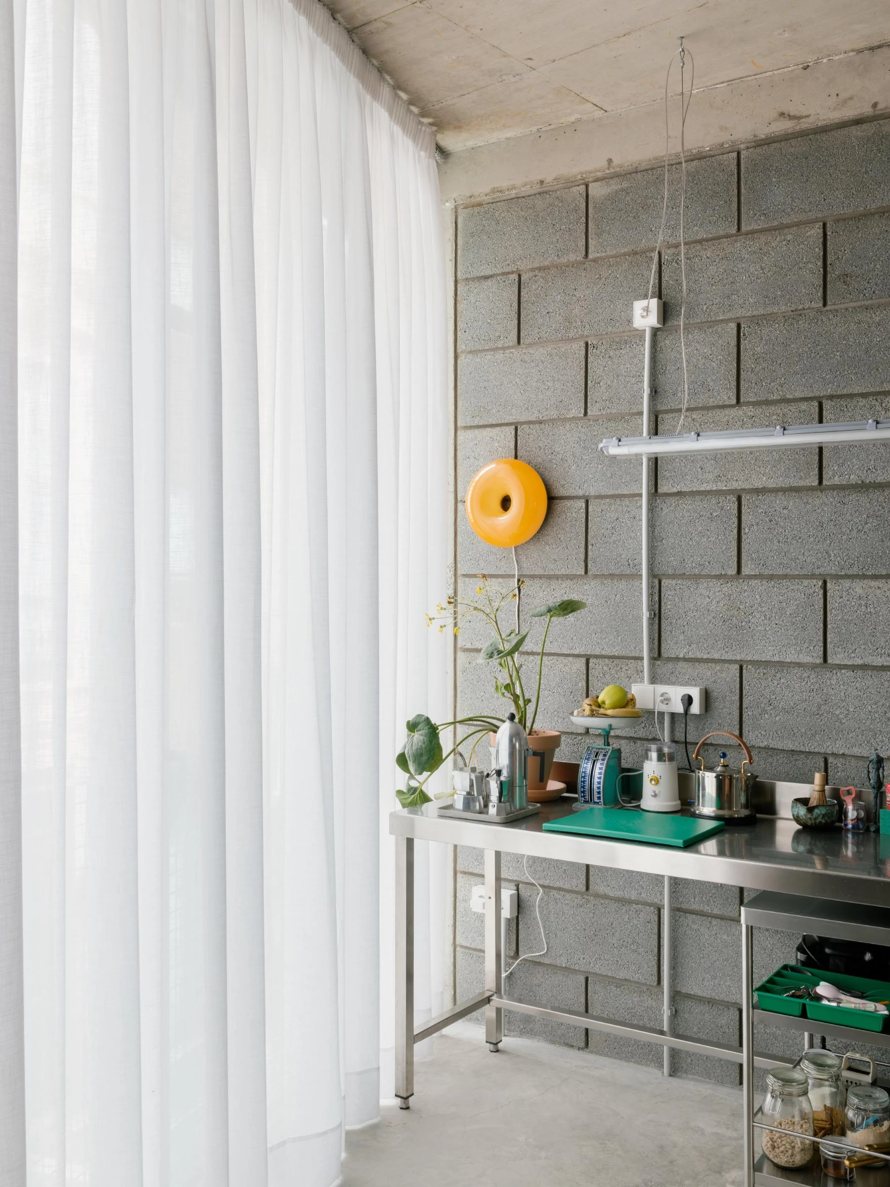 Casa Iriarte by SOCO Estudio, Simone Marcolin: stainless kitchen workbench hung from picon block, a yellow ceramic disc on the wall, sheer linen curtain filtering daylight.