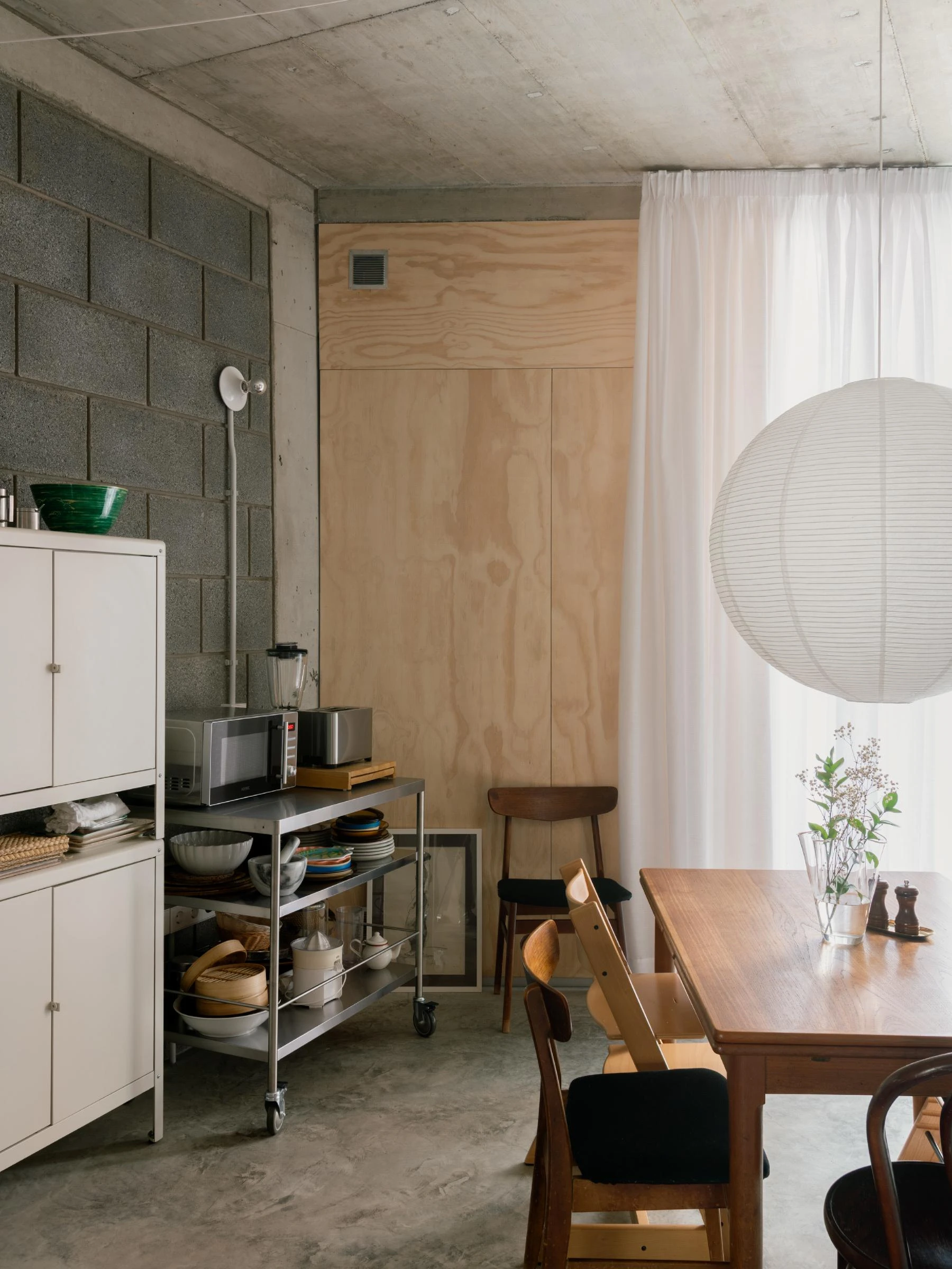 Casa Iriarte by SOCO Estudio, Simone Marcolin: kitchen and dining corner with a stainless trolley island, a white sheer curtain, a paper lantern and a dark timber table set against picon block.