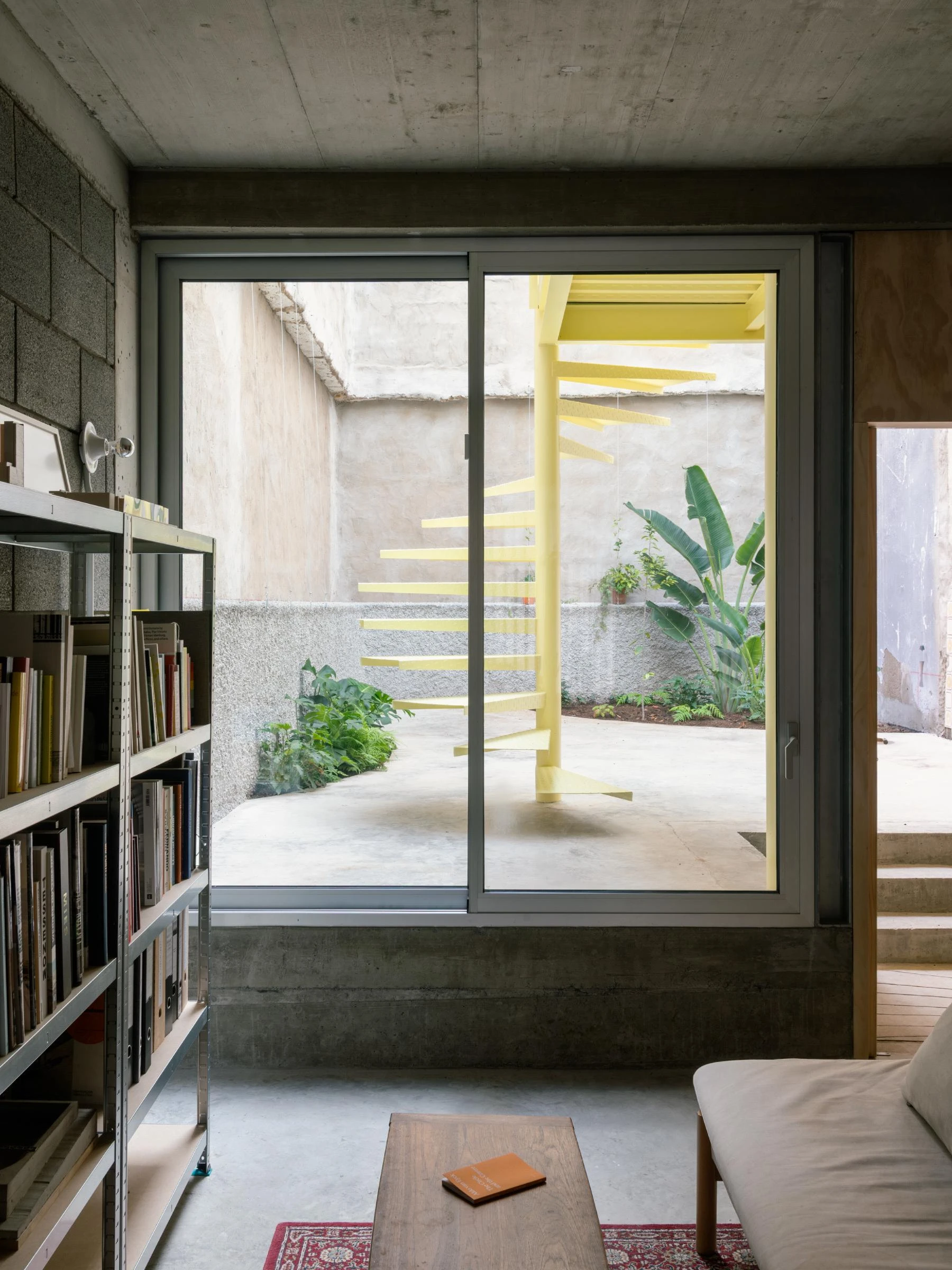 Casa Iriarte by SOCO Estudio, Simone Marcolin: ground-floor library opens onto the courtyard, where the yellow steel spiral stair coils against banana leaves and patinated render.