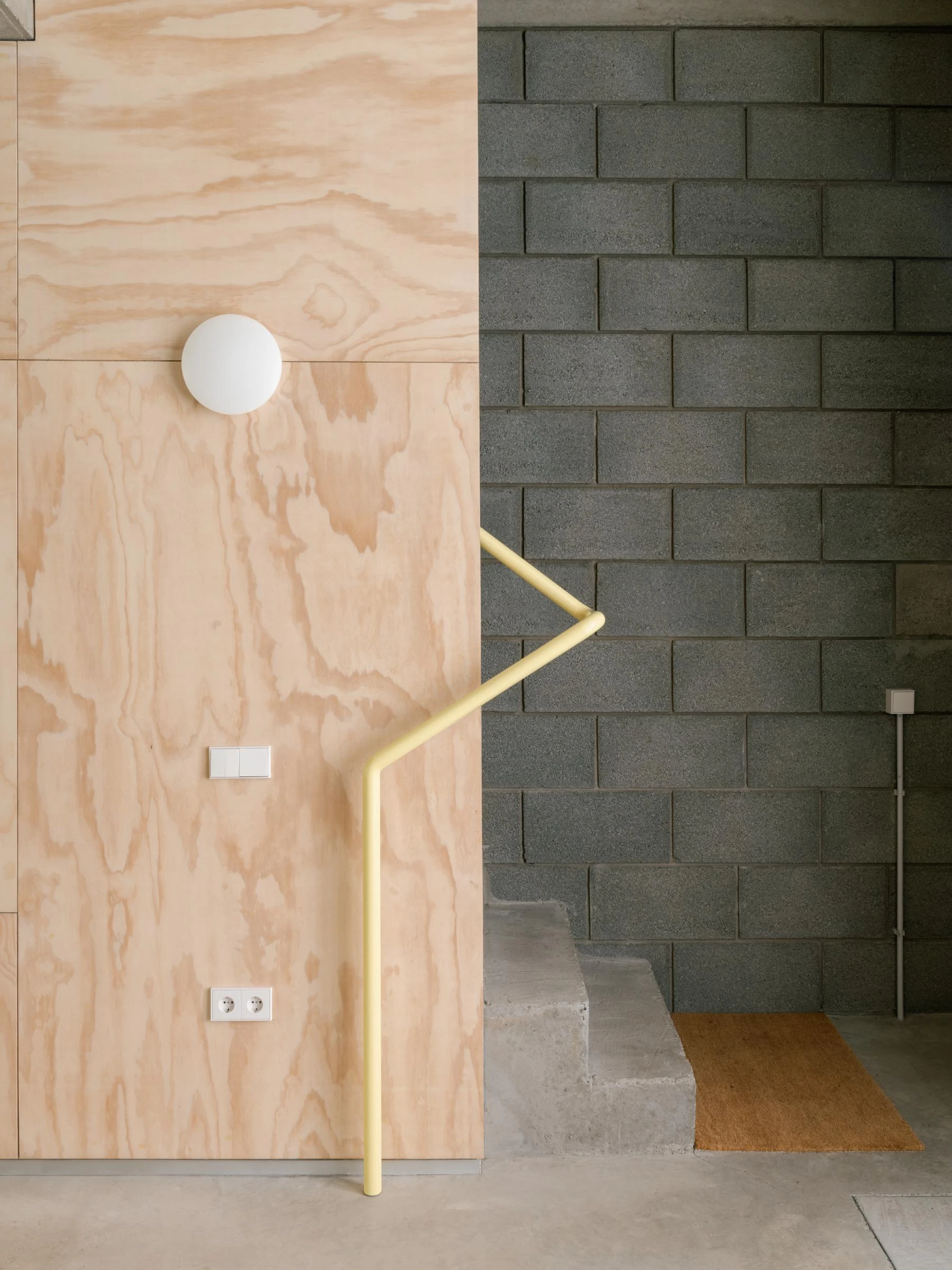 Casa Iriarte by SOCO Estudio, Simone Marcolin: detail at the stair with a yellow powder-coated handrail bent along pine plywood, picon block on the right, a circular wall light reading as a moon.
