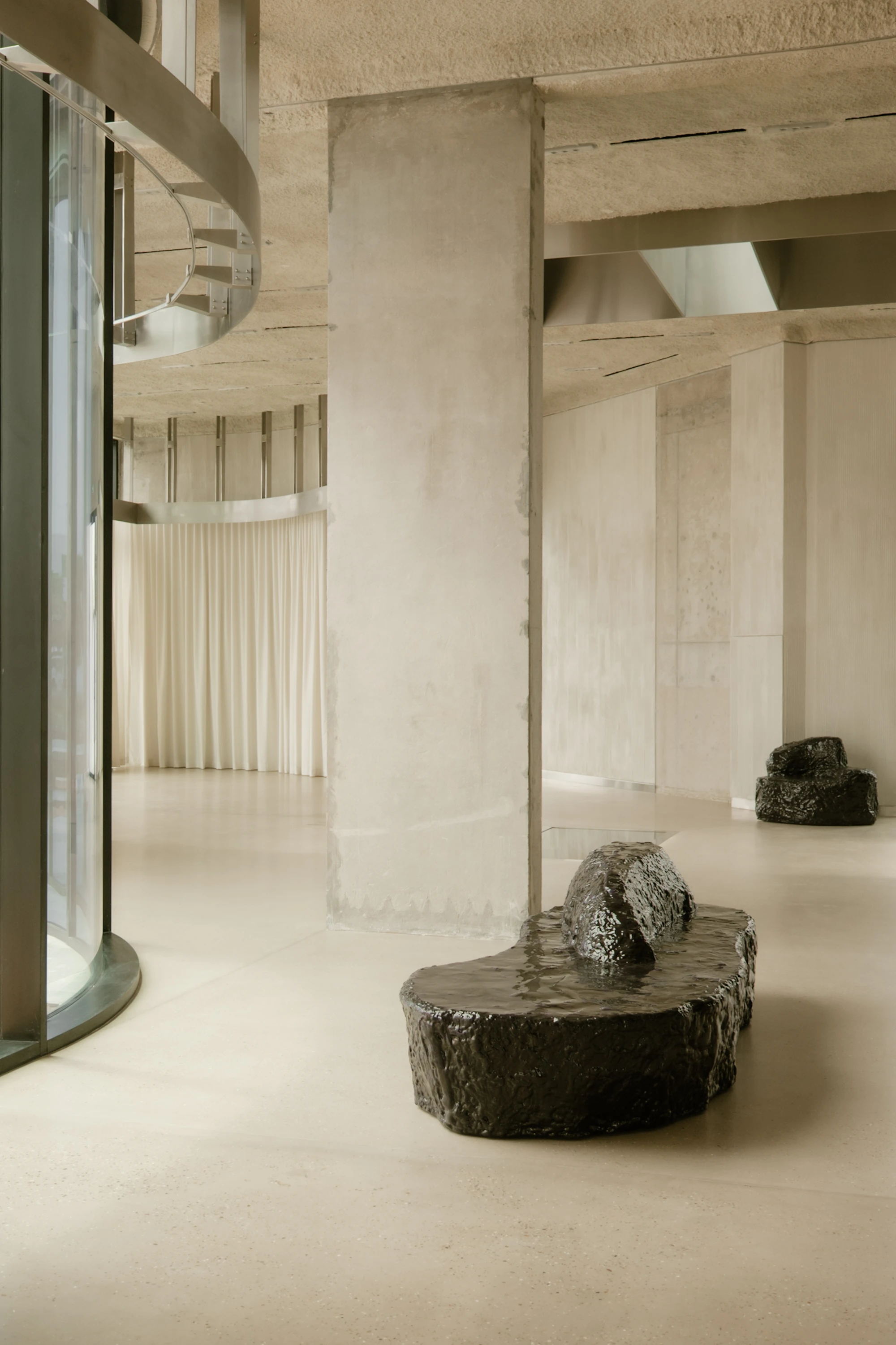 OUNASS STAGE by VAUST, photographed by Nicolas Quiniou — board-formed concrete columns and a curved glass staircase rising from the rear wall, with a Balzer Balzer boulder bench in cast resin on the polished concrete floor.