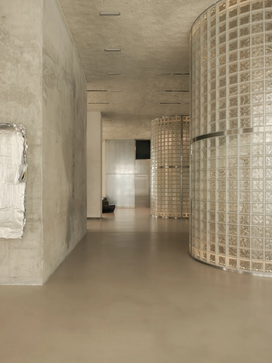 OUNASS STAGE by VAUST, photographed by Nicolas Quiniou — circulation view past a board-formed concrete wall toward a glass-block silo, with a crumpled-foil tapestry mounted to the concrete and ceiling slot reveals lit from above.