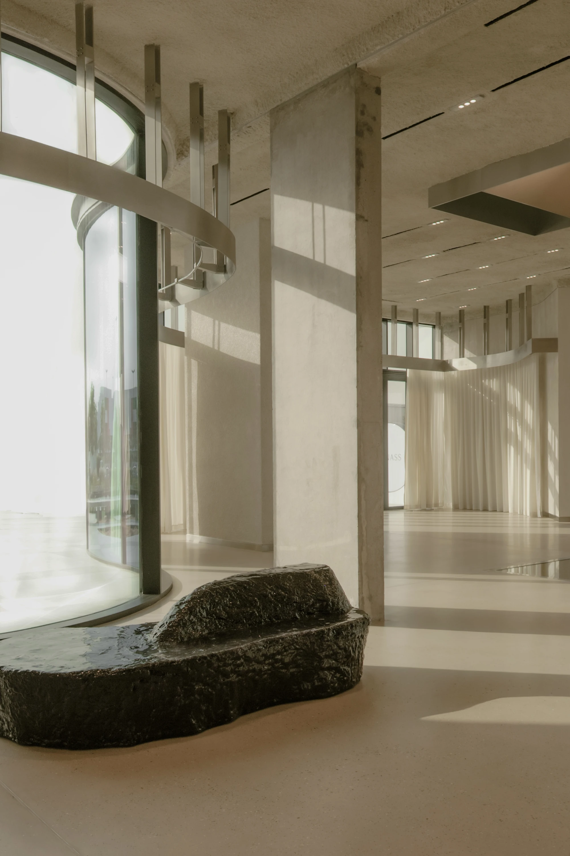 OUNASS STAGE by VAUST, photographed by Nicolas Quiniou — a Balzer Balzer boulder bench in cast resin set in front of a curved floor-to-ceiling window, with sheer white curtains and board-formed concrete columns receding into the deep floorplate.