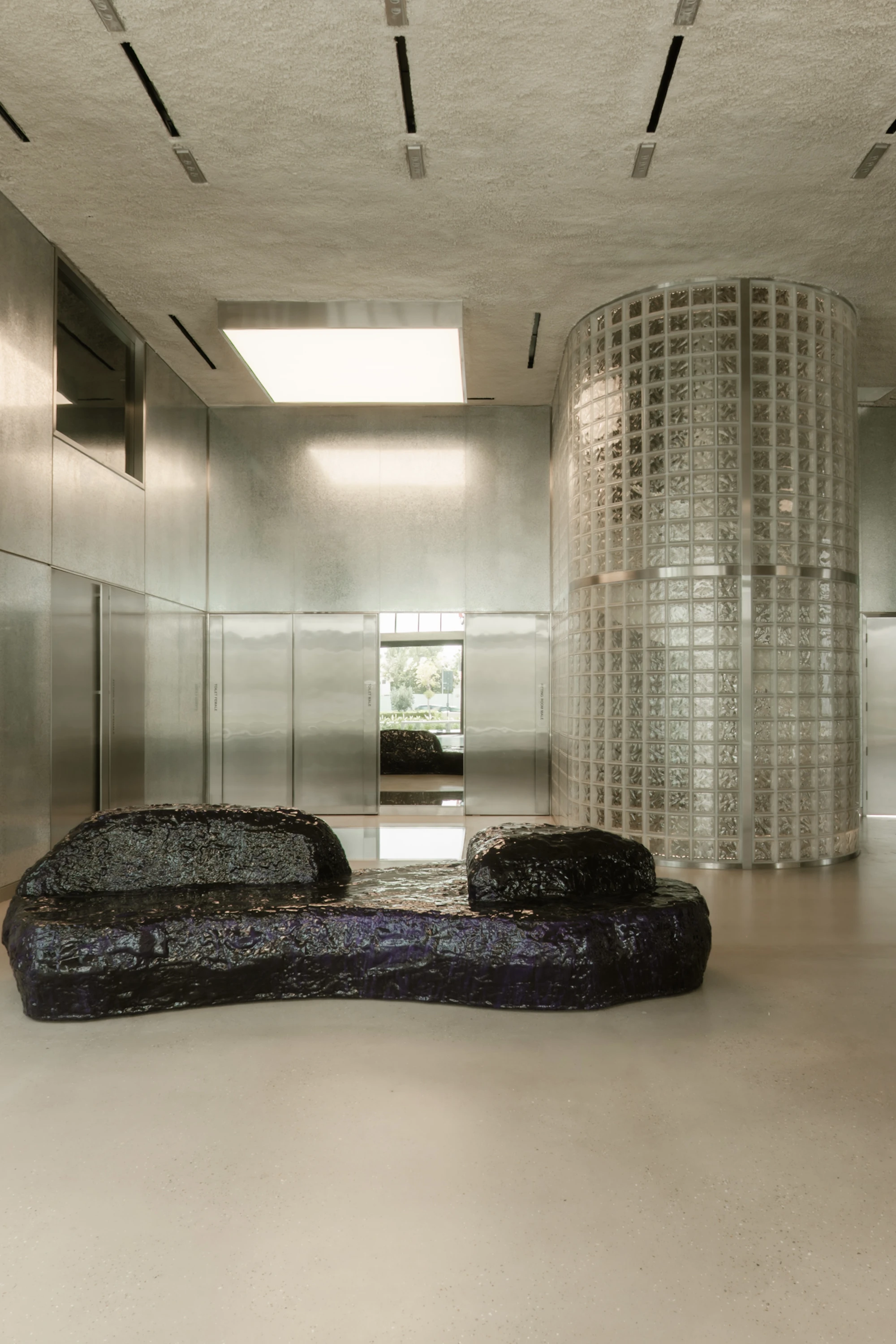 OUNASS STAGE by VAUST, photographed by Nicolas Quiniou — a Balzer Balzer boulder sofa in cast resin set in front of the polished galvanized steel rear wall and the lit glass-block silo housing the changing rooms.