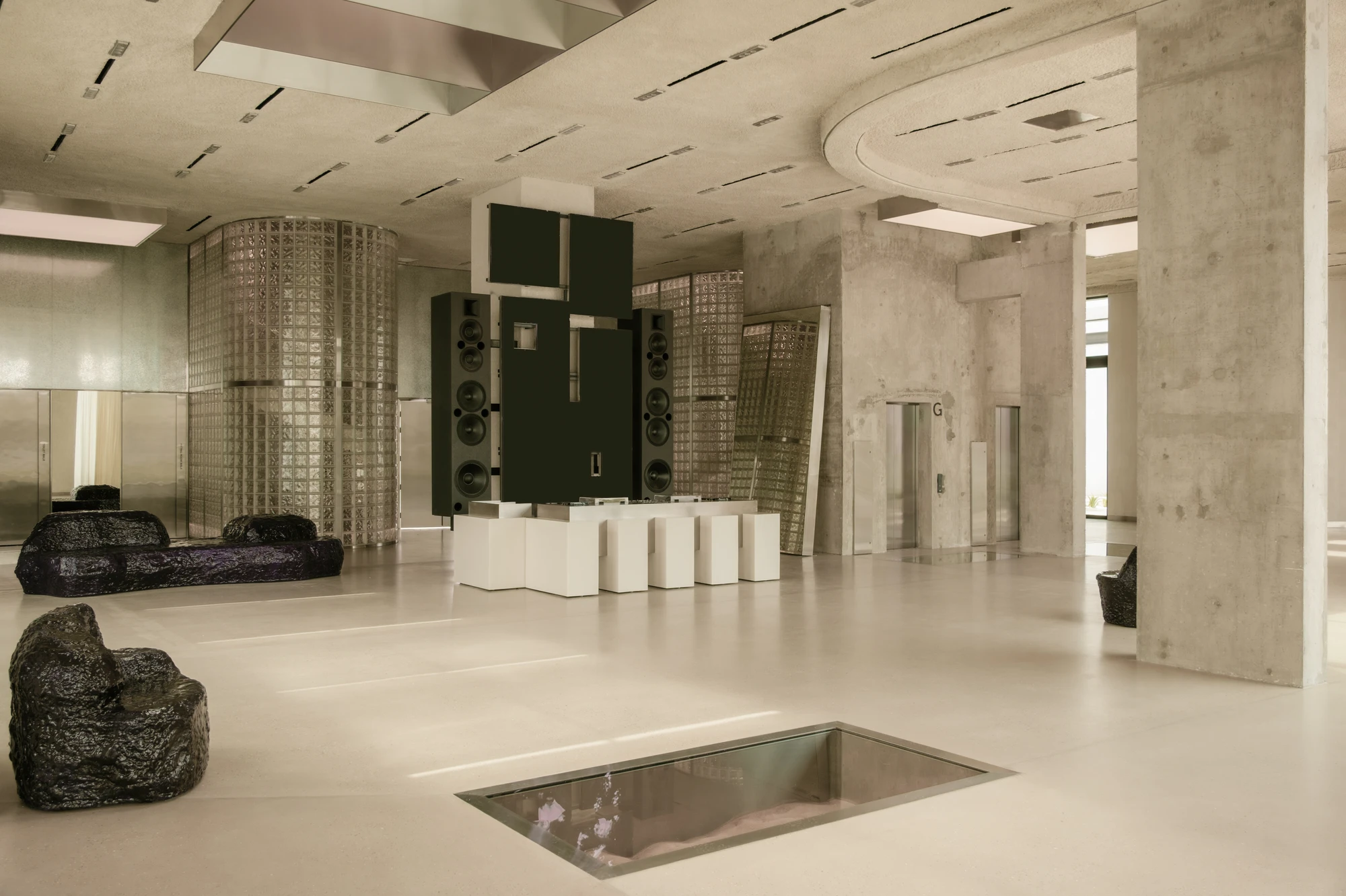 OUNASS STAGE by VAUST, photographed by Nicolas Quiniou — wide axonometric view of the central floor: board-formed concrete columns, the DJ plinth with studio-monitor tower, two glass-block silos, Balzer Balzer boulder seating and a sand pit set into the s