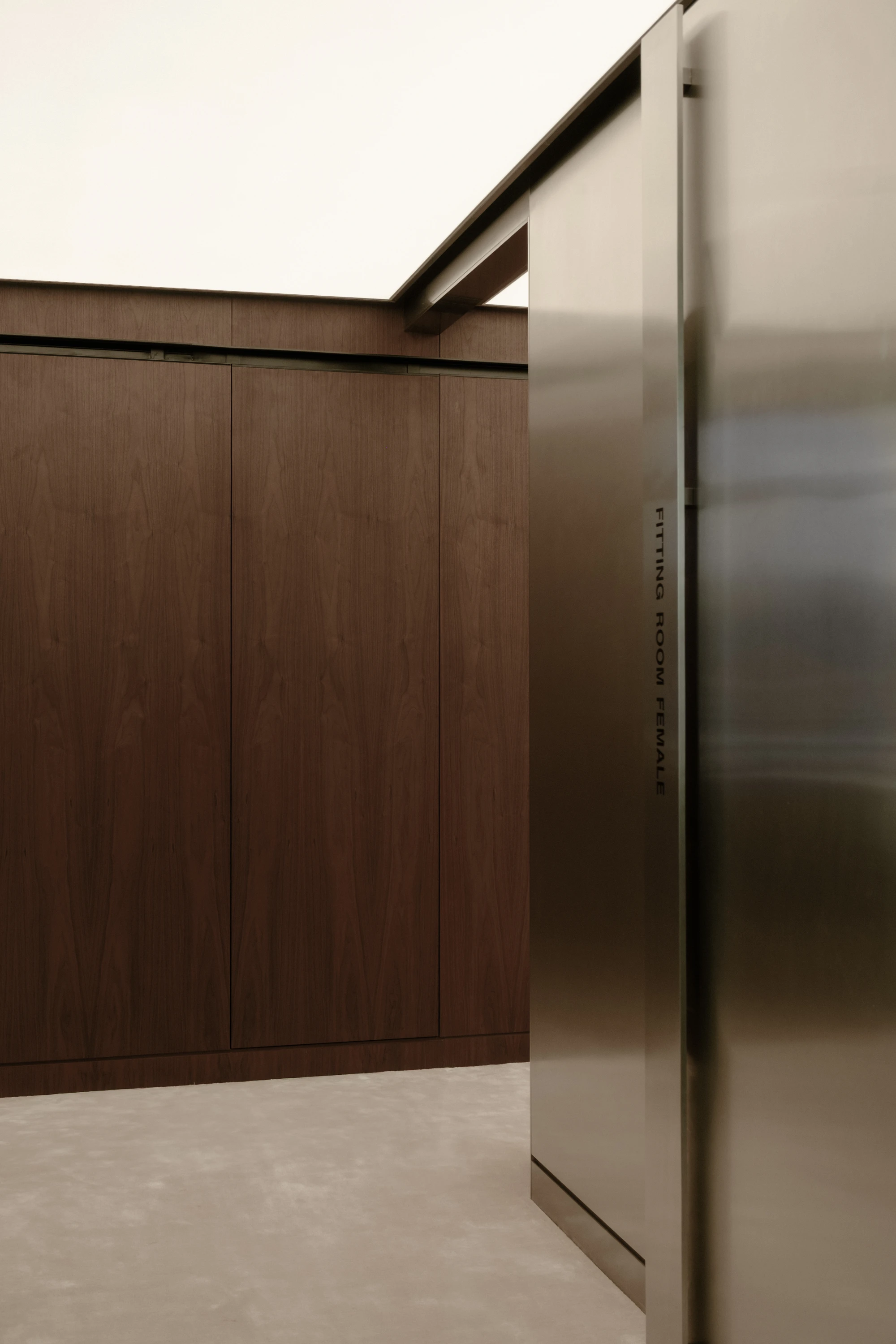 OUNASS STAGE by VAUST, photographed by Nicolas Quiniou — interior detail of a changing-room volume in dark walnut joinery meeting a stainless steel doorframe engraved with the vertical Fitting Room Temporary label.
